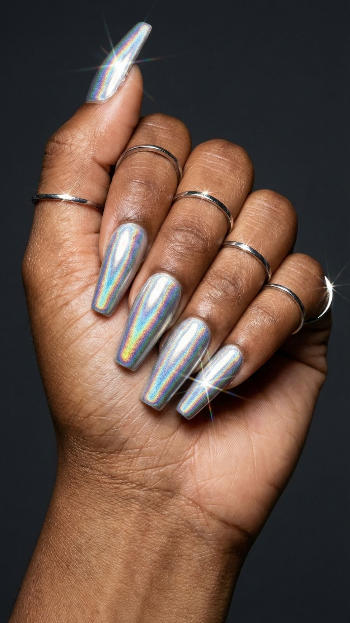 A hand with long, holographic chrome nails, accessorized with several thin silver rings on each finger, shown against a dark background. The shiny Chrome Nails reflect rainbow-colored light, giving this selfie nails look a bold and metallic finish.