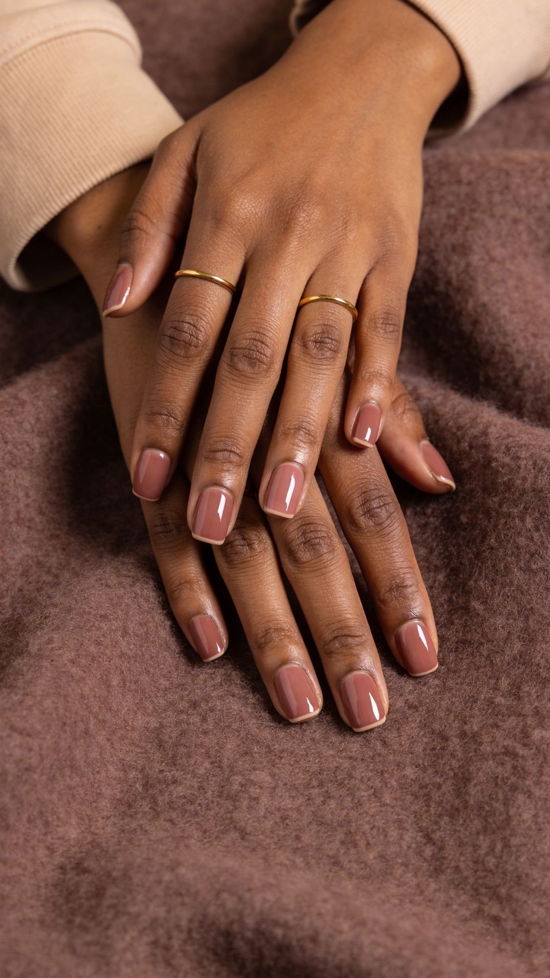 Two hands with neatly manicured, shiny nude-colored nails—perfect wedding guest nails—rest on soft brown fabric. The person wears a beige long-sleeve top and two simple gold rings on their fingers.