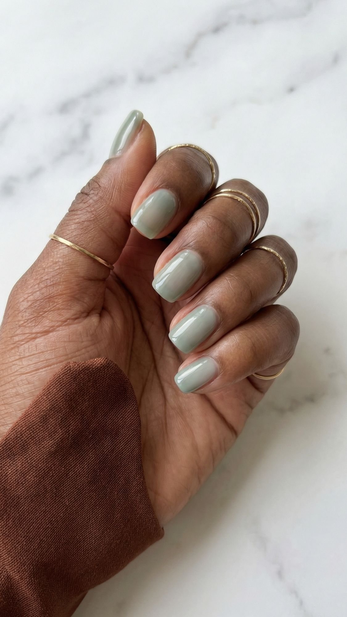 A hand with polished, light green clean girl nails wears several thin gold rings, set against a white marble background. The person is dressed in a brownish long-sleeve top—perfect inspiration for everyday nails.