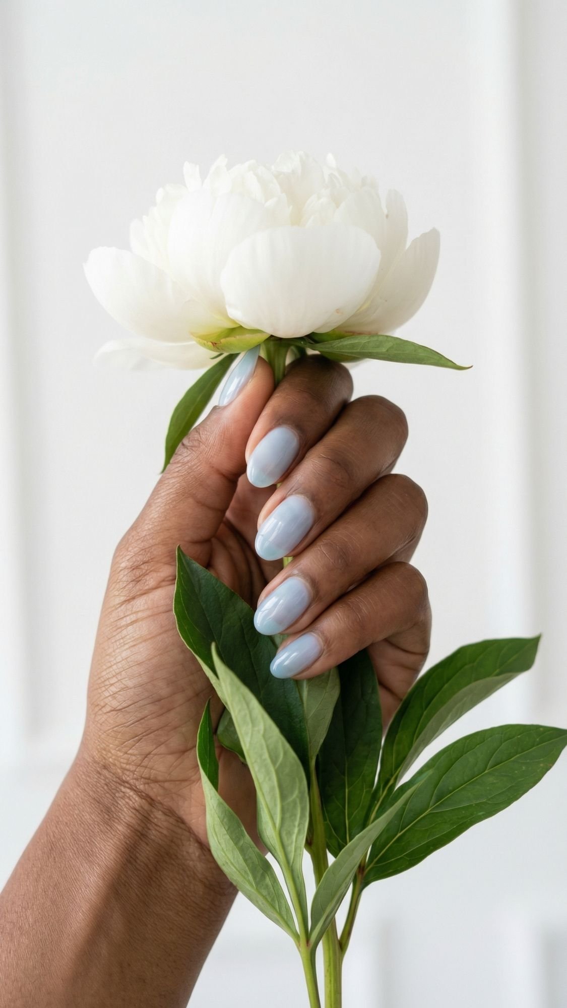 A hand with light blue polished short nails holds a white peony flower with green leaves against a soft, white background, showcasing elegant nails.