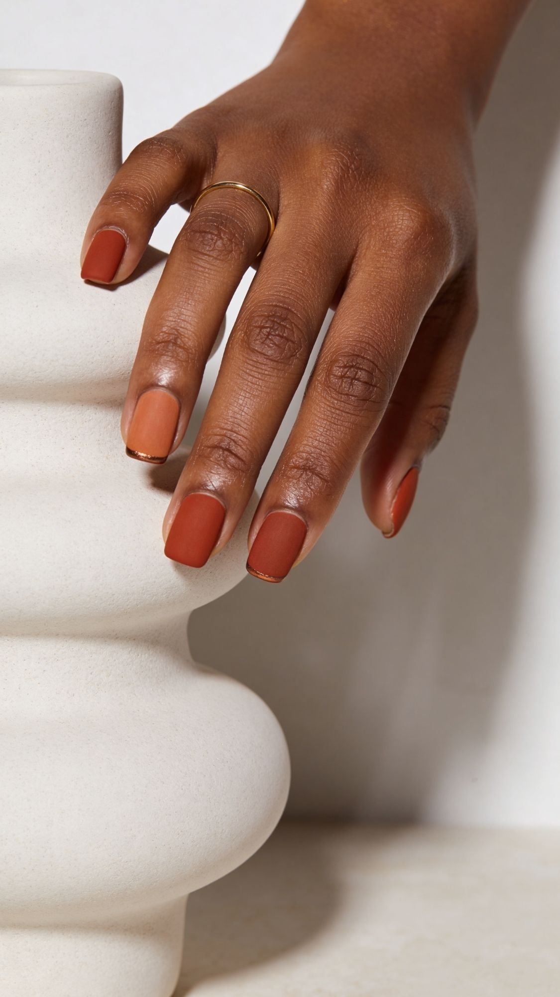 A hand with smooth brown skin and neatly manicured brunch nails painted in a matte terracotta shade rests gently on a white, sculptural ceramic vase. A thin gold ring adorns the ring finger.