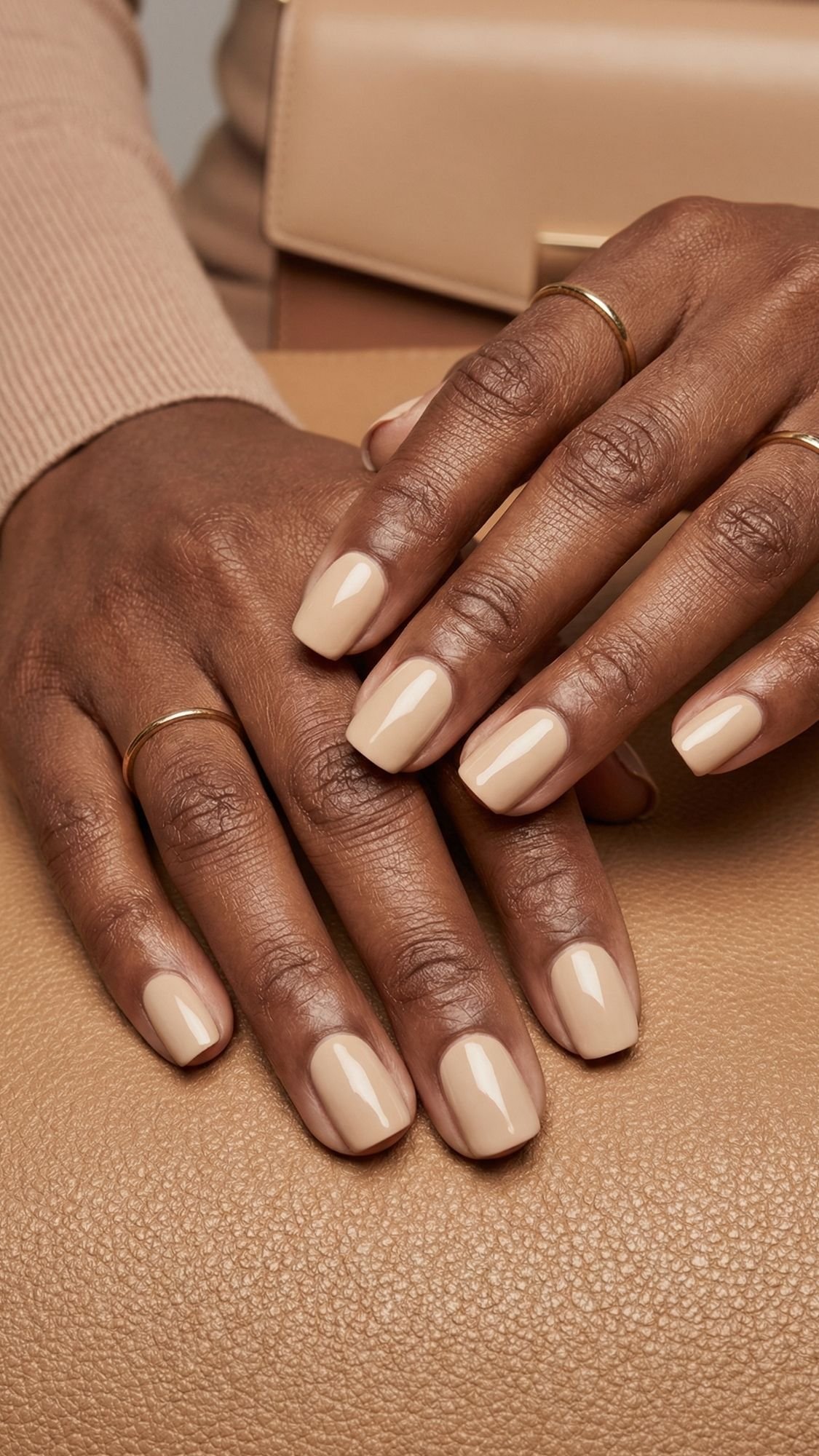 Close-up of hands with short, neatly manicured nails painted in a glossy nude beige polish—perfect wedding guest nails. The person wears thin gold rings, with tan and beige textures from a purse and sweater in the background.