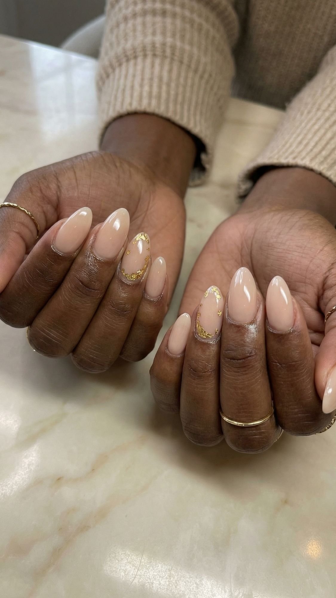 Two hands with flattering almond nails in a nude shade, featuring gold foil accents, rest on a marble surface. The person wears gold rings and a beige knit sweater.