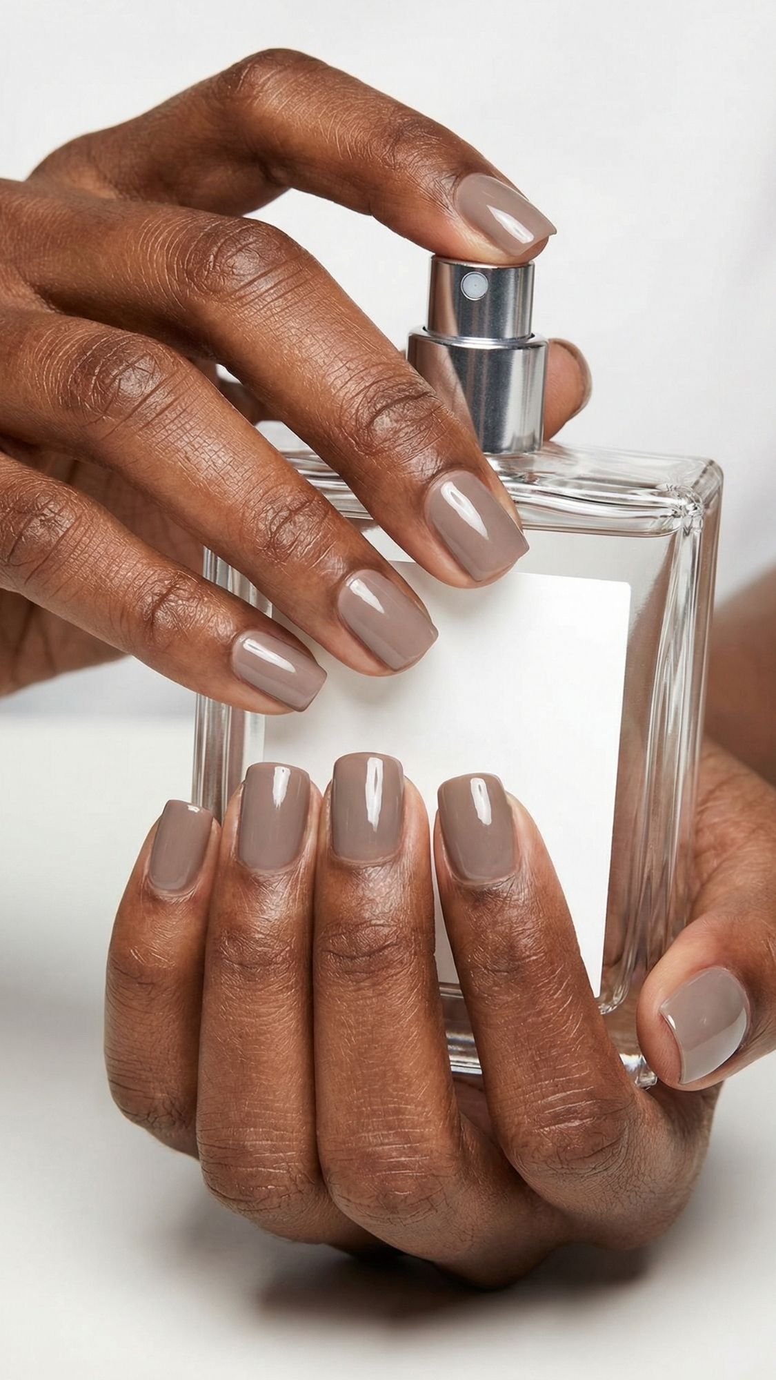 A person with neatly manicured Clean Girl Nails painted in taupe holds a clear glass perfume bottle with a silver spray nozzle against a plain background, showcasing the perfect Polished Everyday Look.