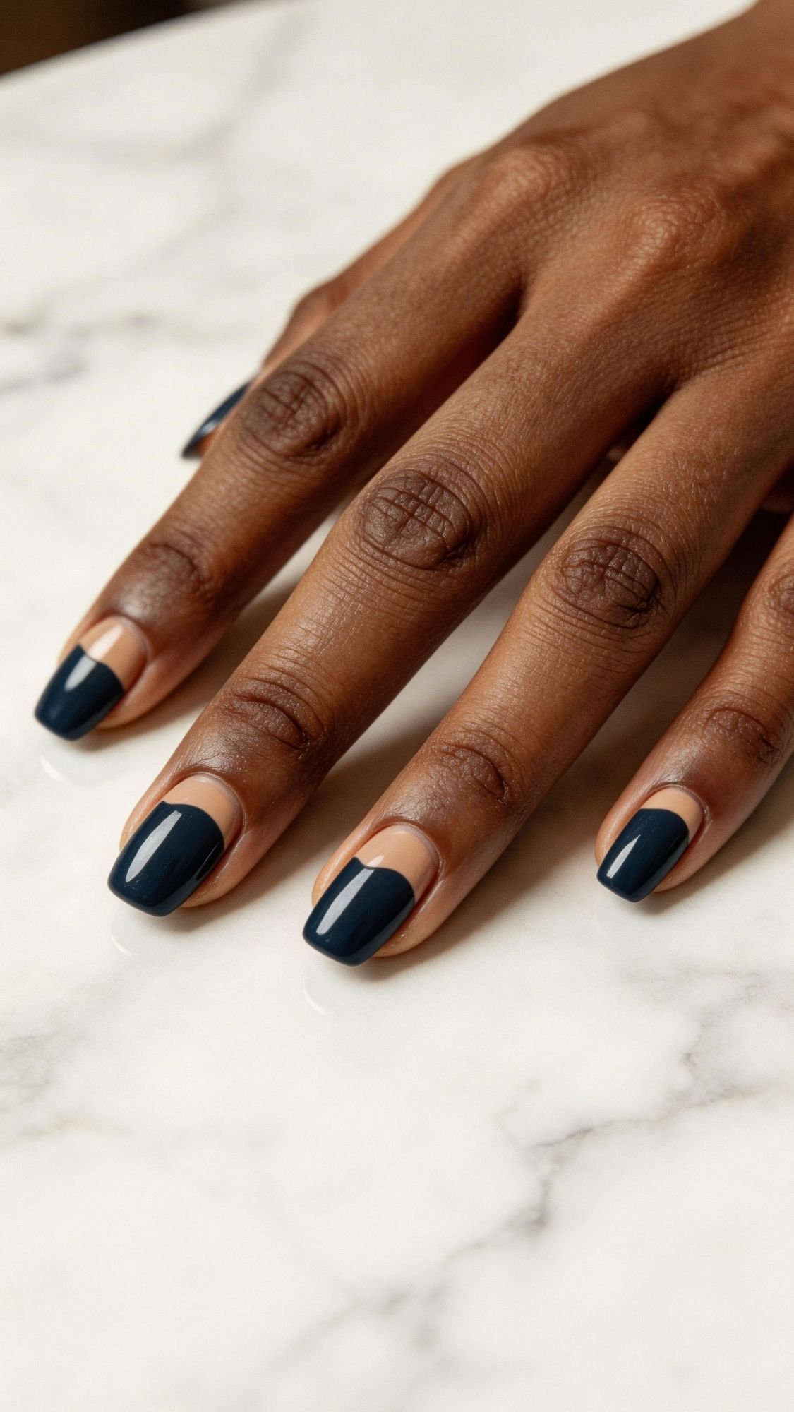 A close-up of a hand with dark skin resting on a marble surface, featuring elegant nails painted with modern dark blue polish and a nude crescent near the cuticle in a chic half-moon nail design on short nails.