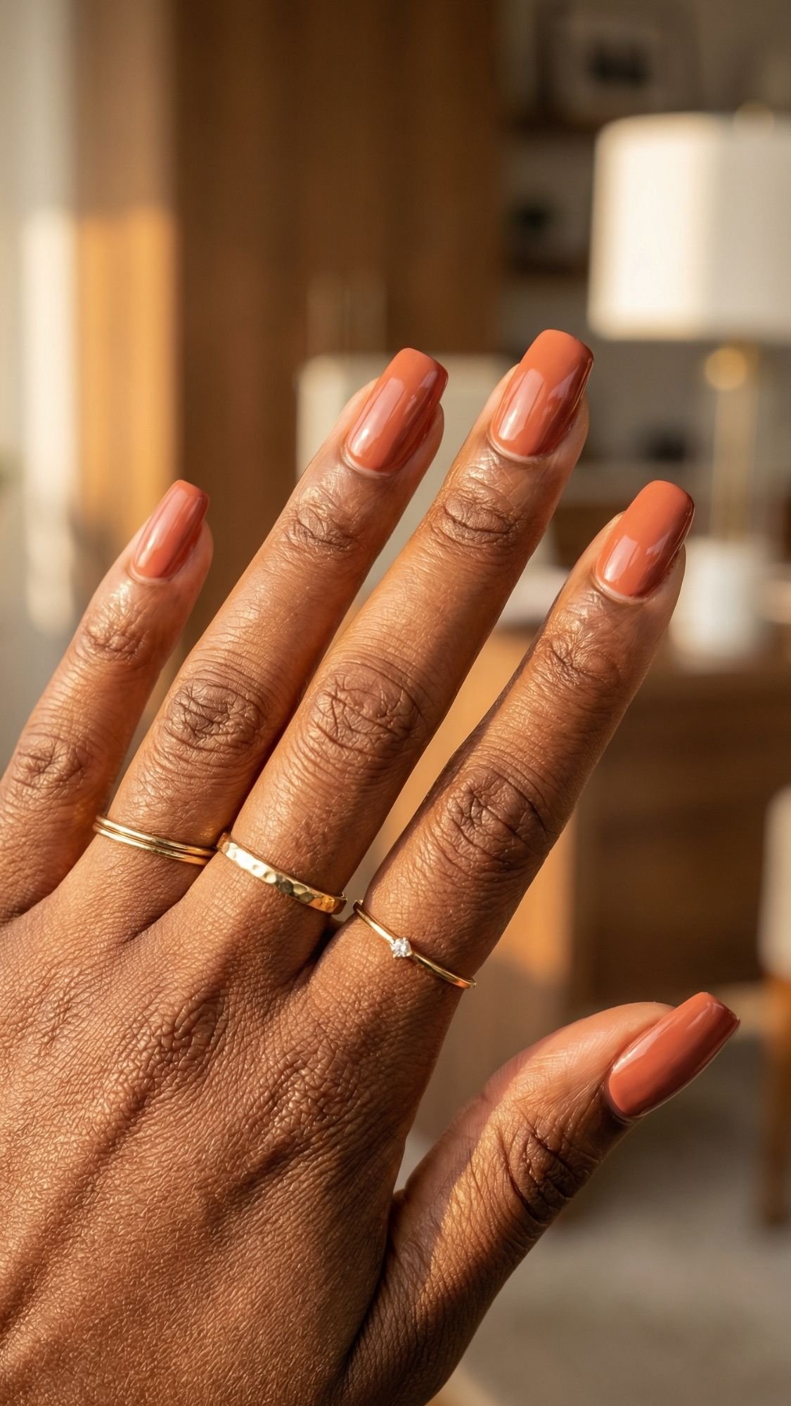 A hand with medium brown skin shows long, square-shaped chic nails painted in a glossy terracotta color. The person wears three delicate gold rings. A blurred wooden desk and lamp are in the background, perfect for work friendly nails.