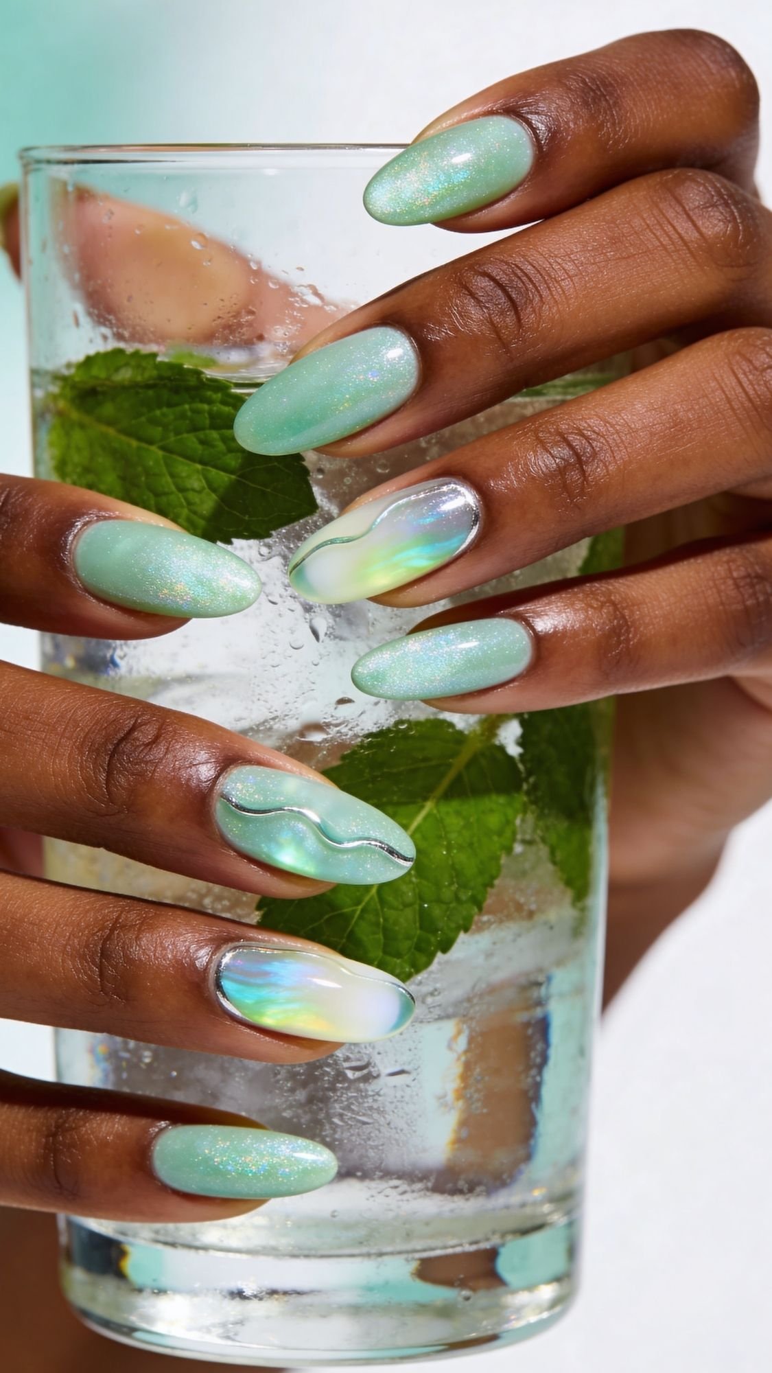 A hand with long, almond-shaped summer nails painted in iridescent mint green and holographic designs holds a glass of sparkling water with mint leaves and ice.