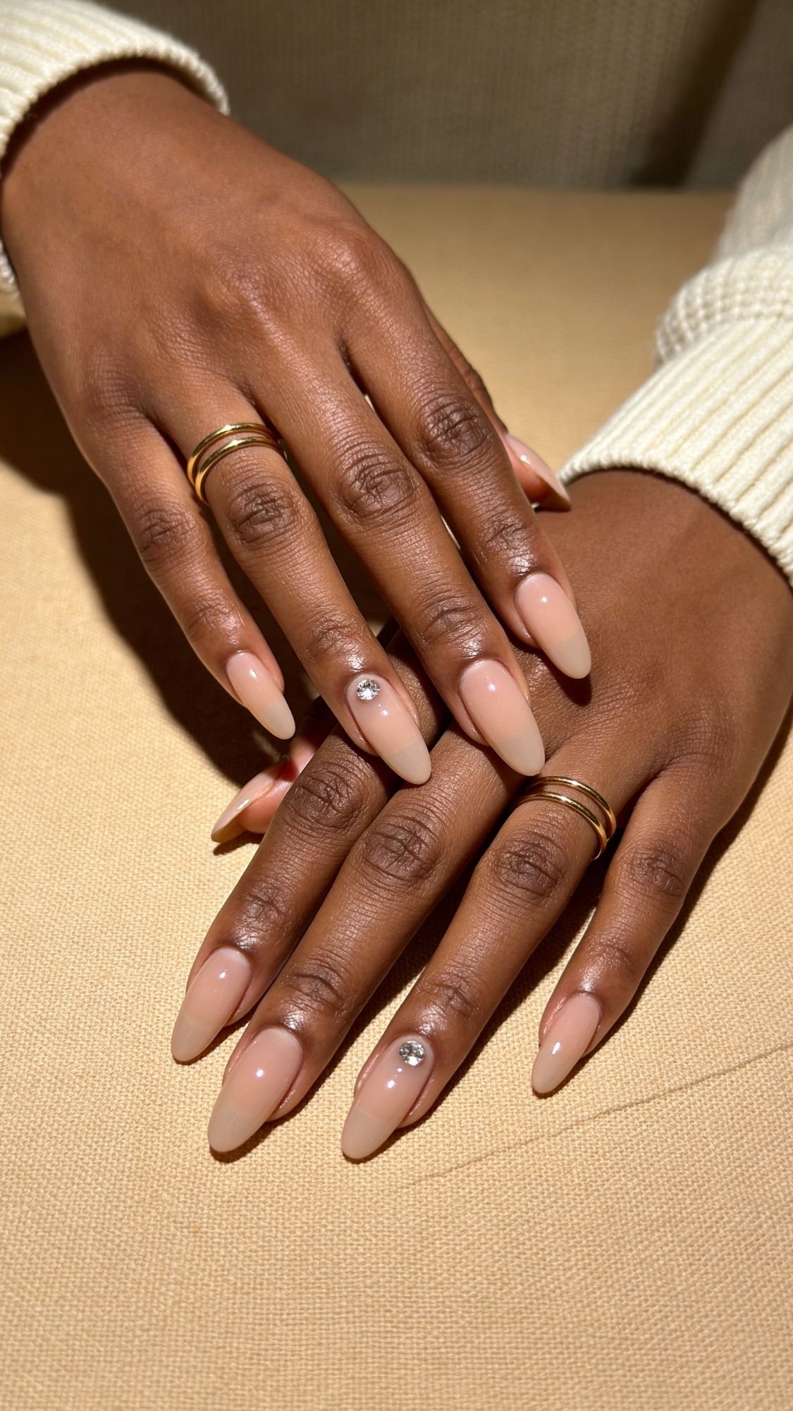 A close-up of two hands with long, almond nails showcases a chic nude Nail Design, each featuring a single rhinestone accent. The person is wearing gold rings on several fingers and a light-colored sweater.