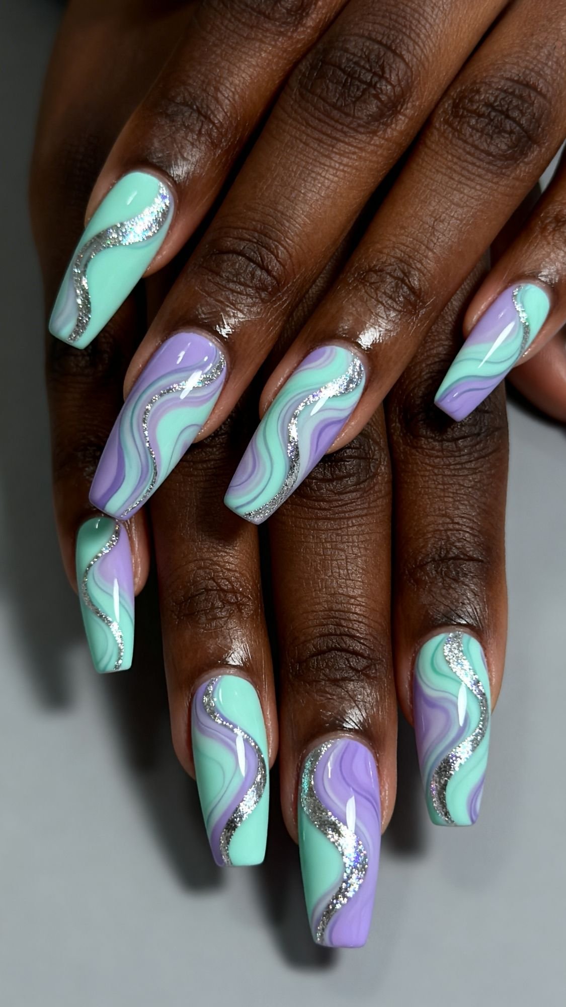 Close-up of hands with long nails featuring a swirl design in mint green and lavender, accented with silver glitter lines for that perfect Sparkle Nails look. Ideal for Easter Holiday Nails against deep brown skin and a neutral gray background.