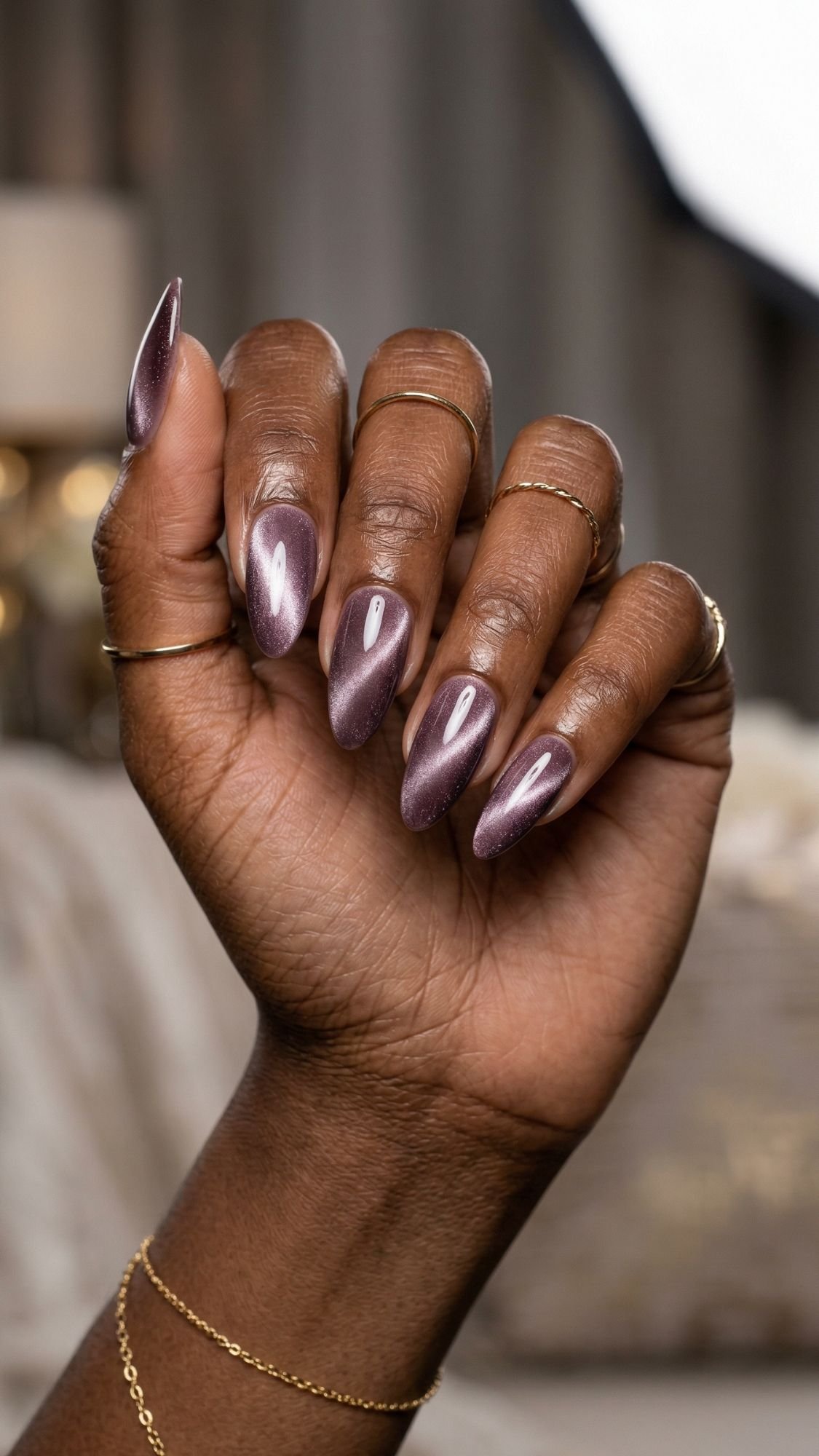 A close-up of a hand with long, almond-shaped chic nails painted in shimmery mauve polish. The person wears delicate gold rings and a bracelet, showcasing a professional nail design against a softly blurred background.