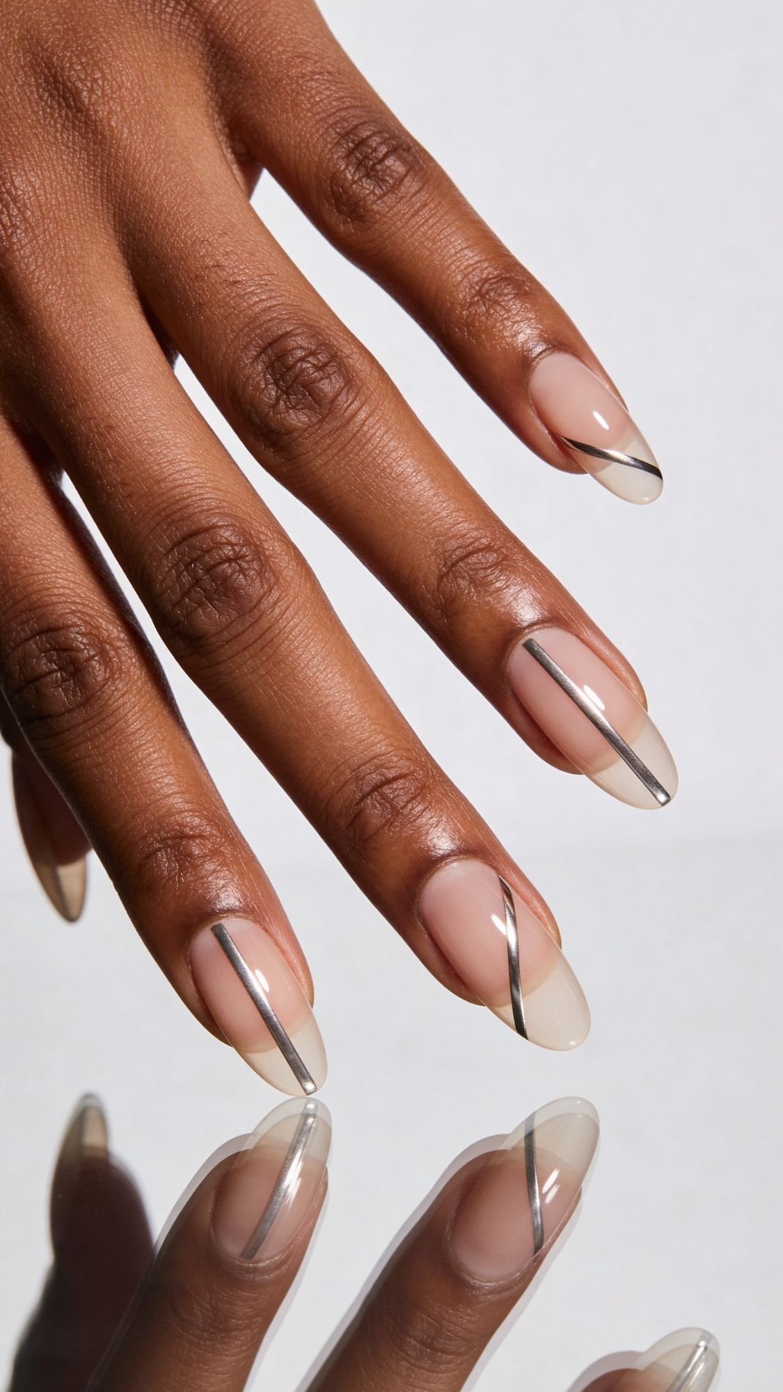 A hand with almond-shaped, light-catching nails features a minimalist manicure: clear polish with thin, angled metallic silver lines on each nail, shown against a light background.
