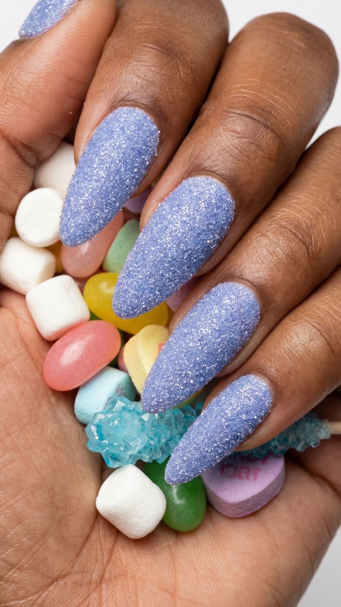 A hand with long, pointed, sparkling nails in glittery lavender holds an assortment of colorful candies, including marshmallows, jelly beans, and rock candy.