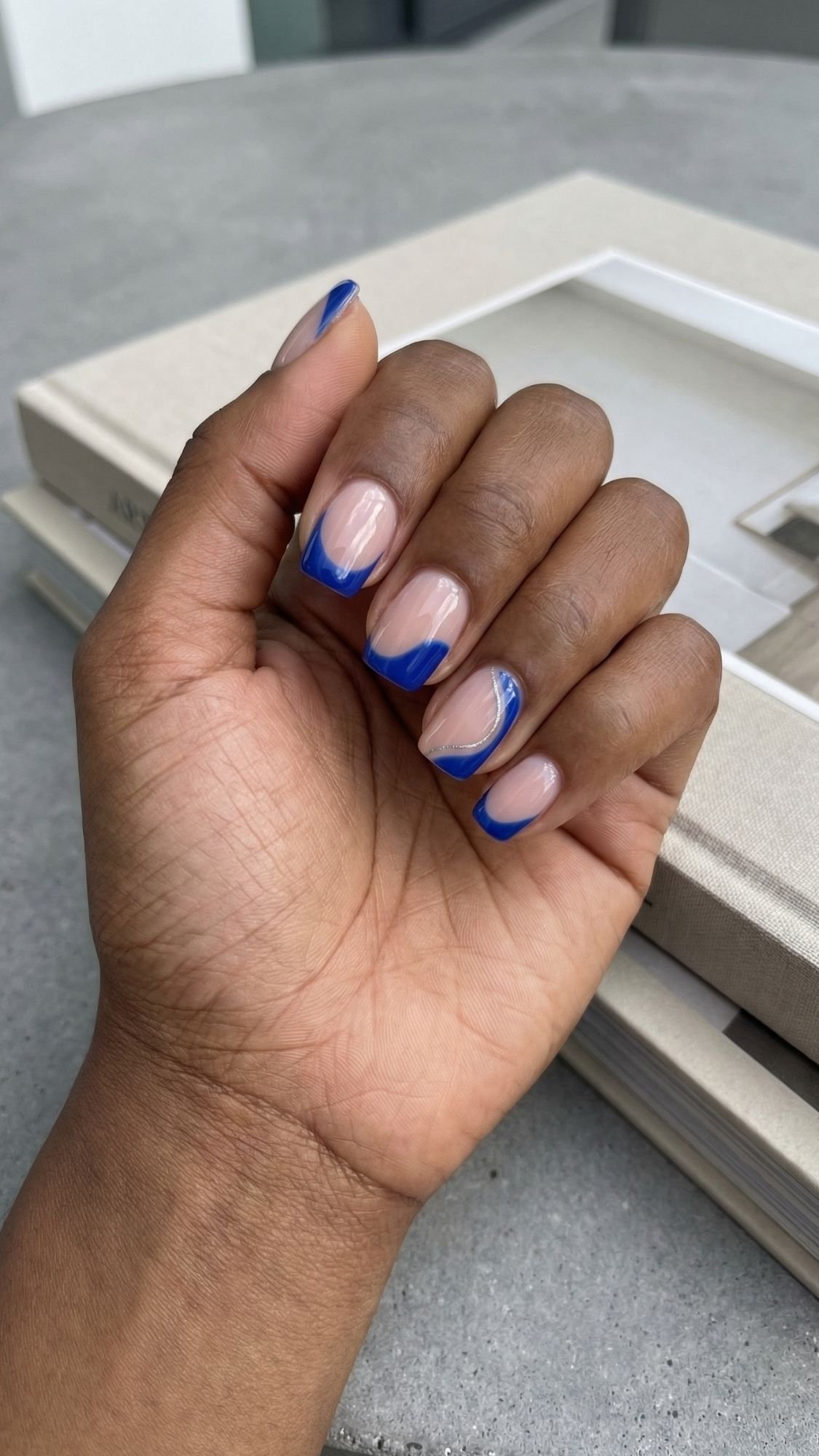 A hand with neatly manicured nails featuring a nude base and bold blue french tips—perfect for Summer Nails—rests on two stacked, beige hardcover books.