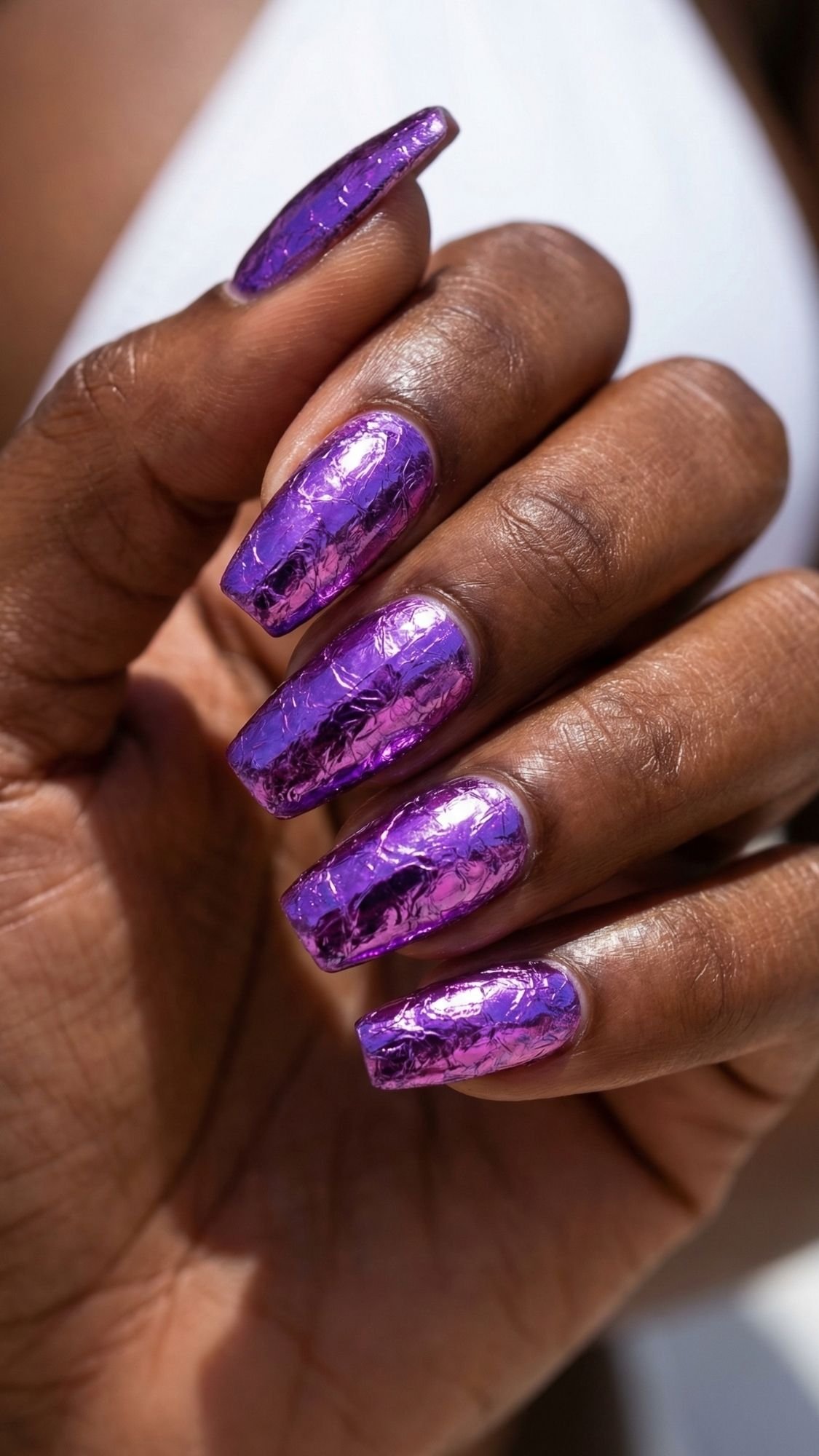 A hand with long, manicured nails painted metallic purple with a marble or foil effect—perfect summer nails—held against a blurred light background.