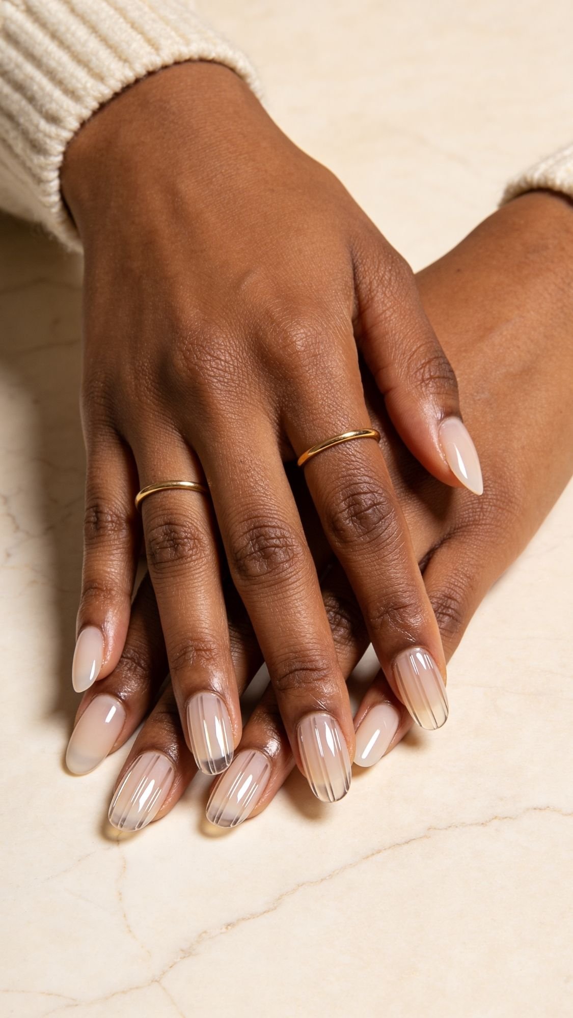 A close-up of hands with almond nails in a nude shade, some featuring fine black vertical lines as chic nail designs. The person is wearing two thin gold rings, and a cream-colored, ribbed sweater sleeve is visible.