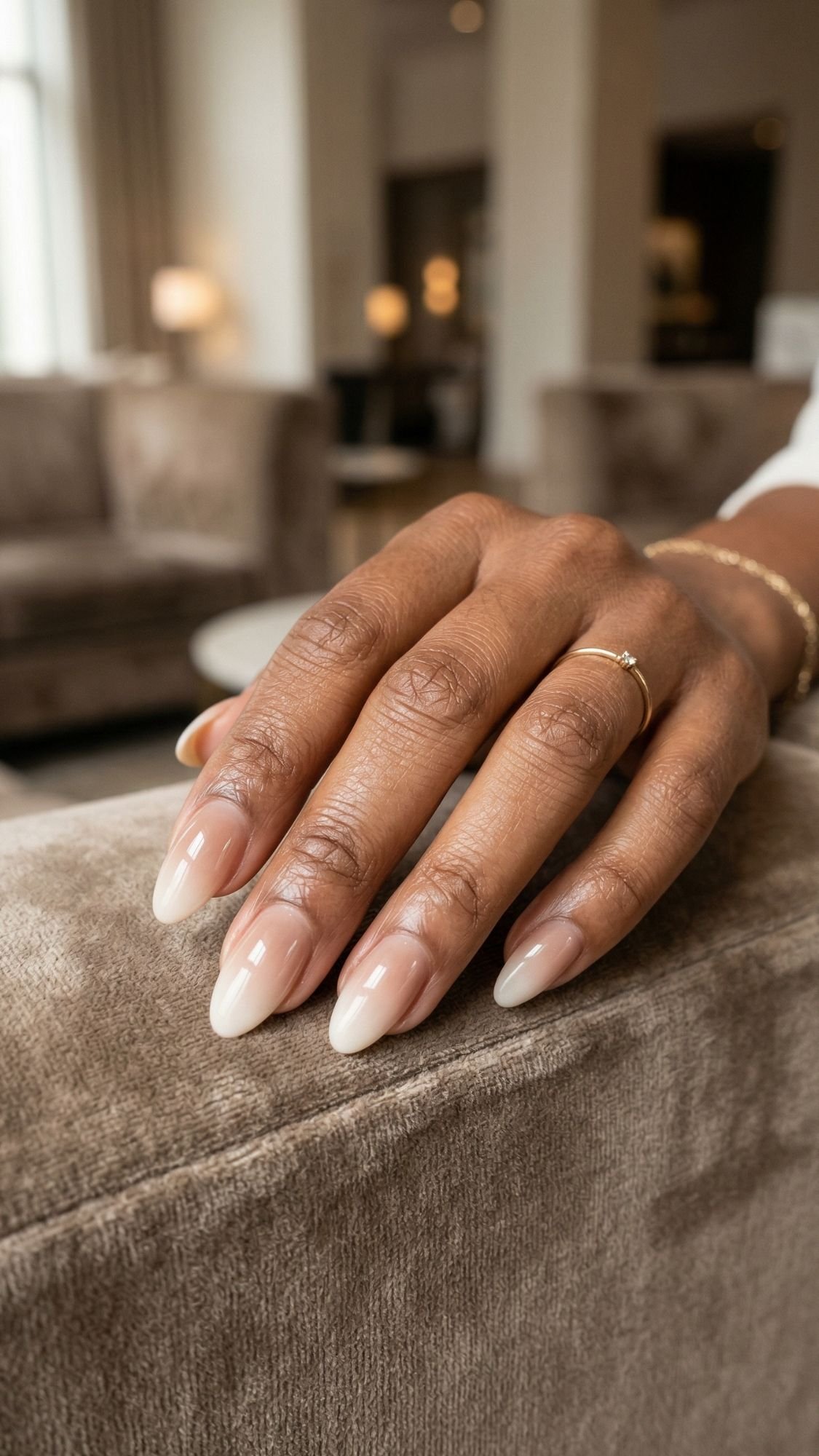 A close-up of a hand with almond-shaped, polished nails painted in a natural, glossy nude color—perfect for an everyday look—resting on a light brown upholstered surface. The person wears a thin gold ring and a simple bracelet against a softly lit living room.