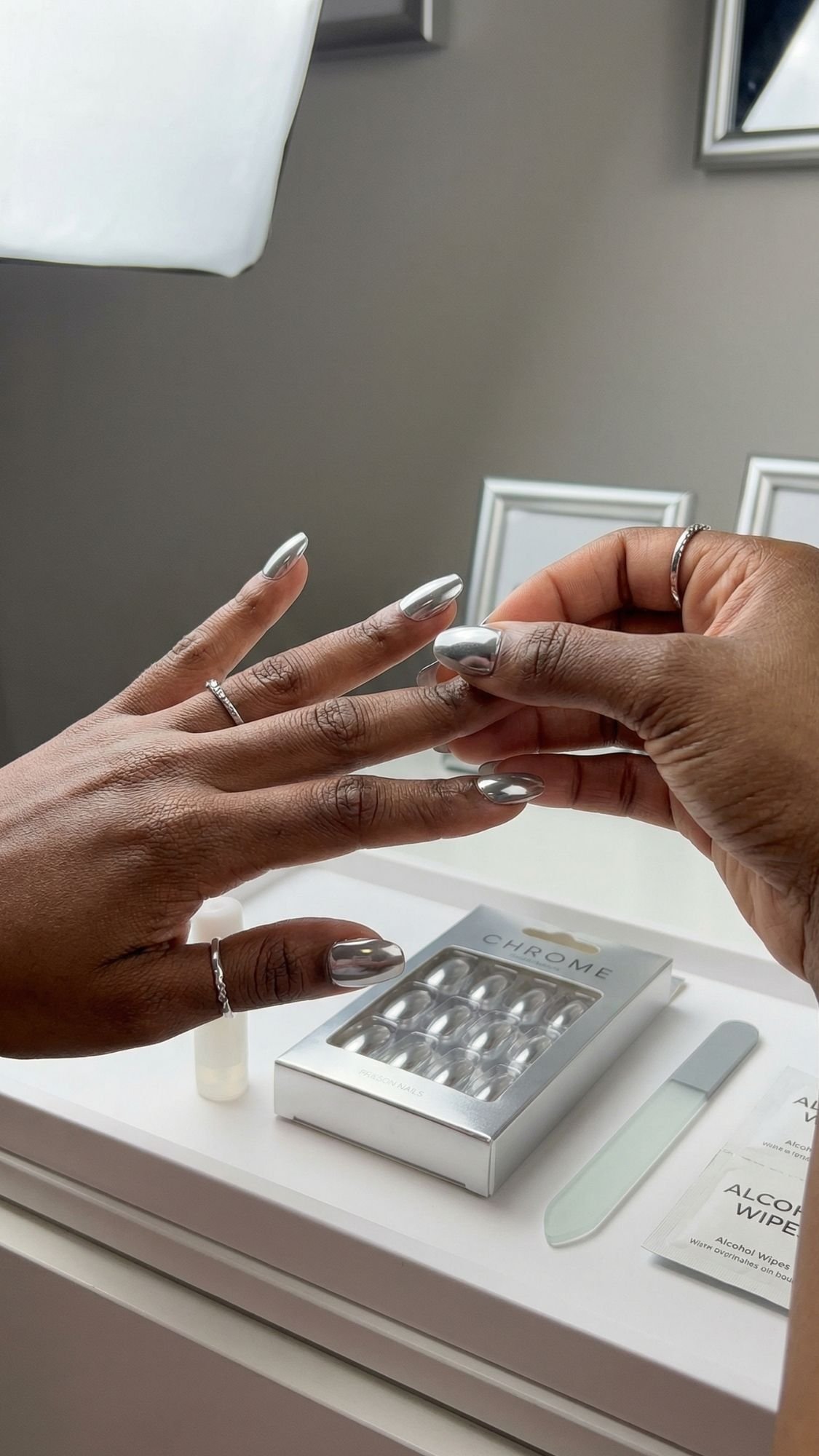 A person applies a silver chrome press-on nail to their left hand, ready for the perfect selfie. On the white table are a box of chrome nails, a nail file, and an alcohol wipe.