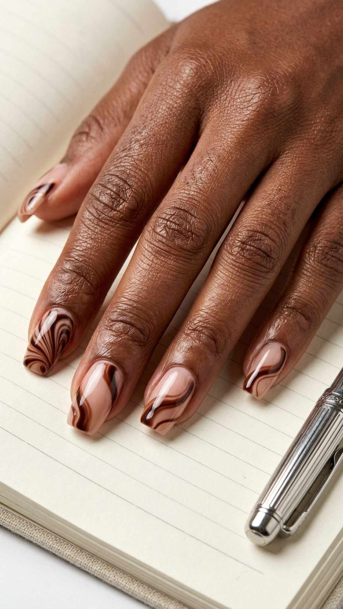 A hand with brown skin rests on an open lined notebook; the nails have a glossy marble design in shades of brown and nude, showcasing chic, work friendly nails. A silver pen lies nearby.