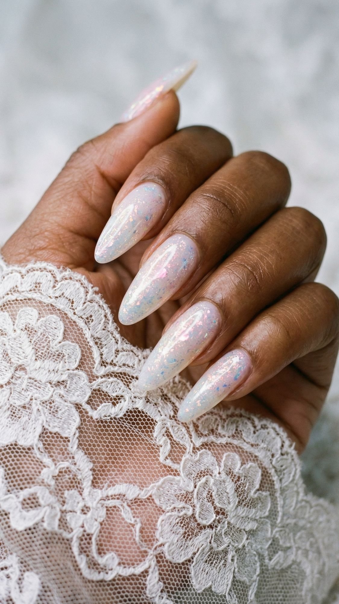 A hand with long, almond-shaped nails painted in pearly, iridescent polish sparkles beautifully as it rests on delicate white lace—perfect for an elegant occasion or Easter Holiday Nails.