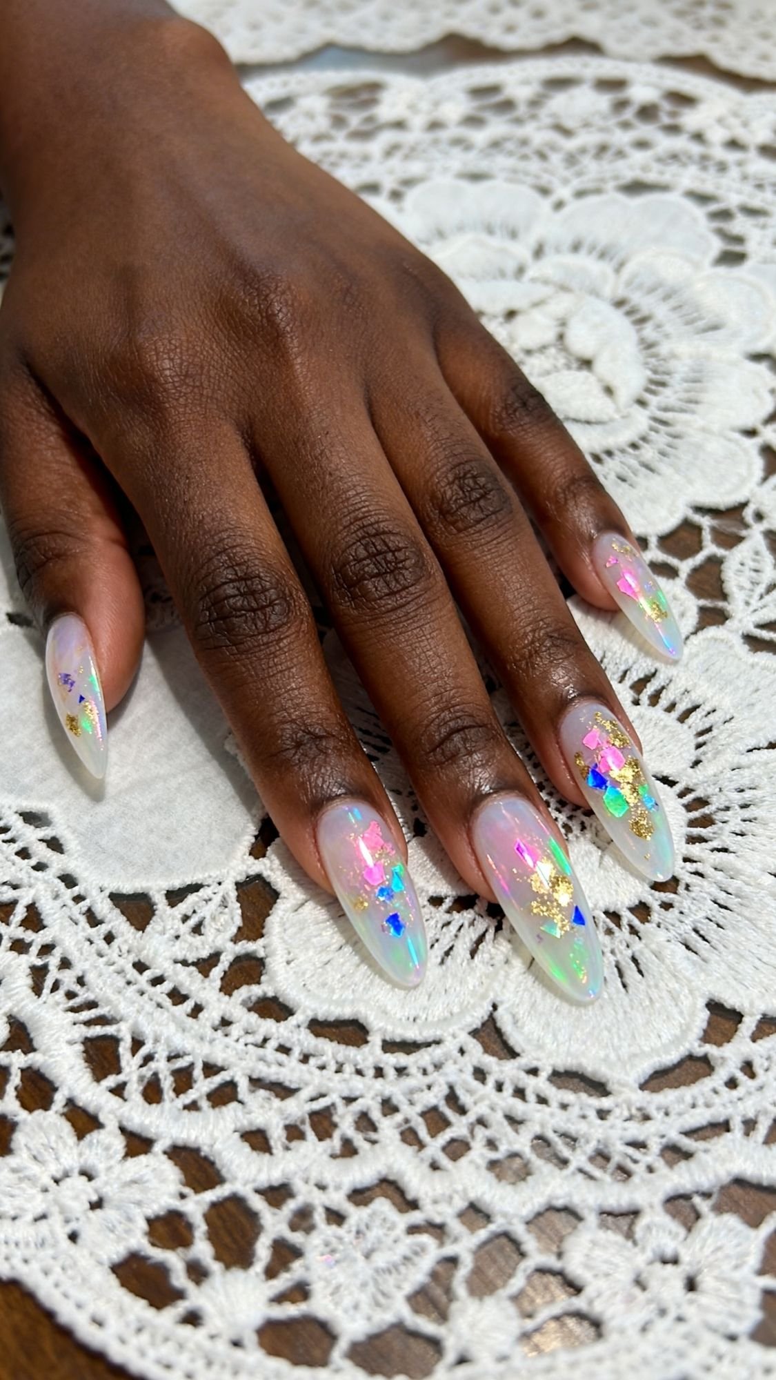A hand with long, almond-shaped sparkle nails featuring iridescent confetti and gold foil designs rests on a white lace doily. The glossy, translucent base and reflective decorations make them perfect for holiday nail designs.