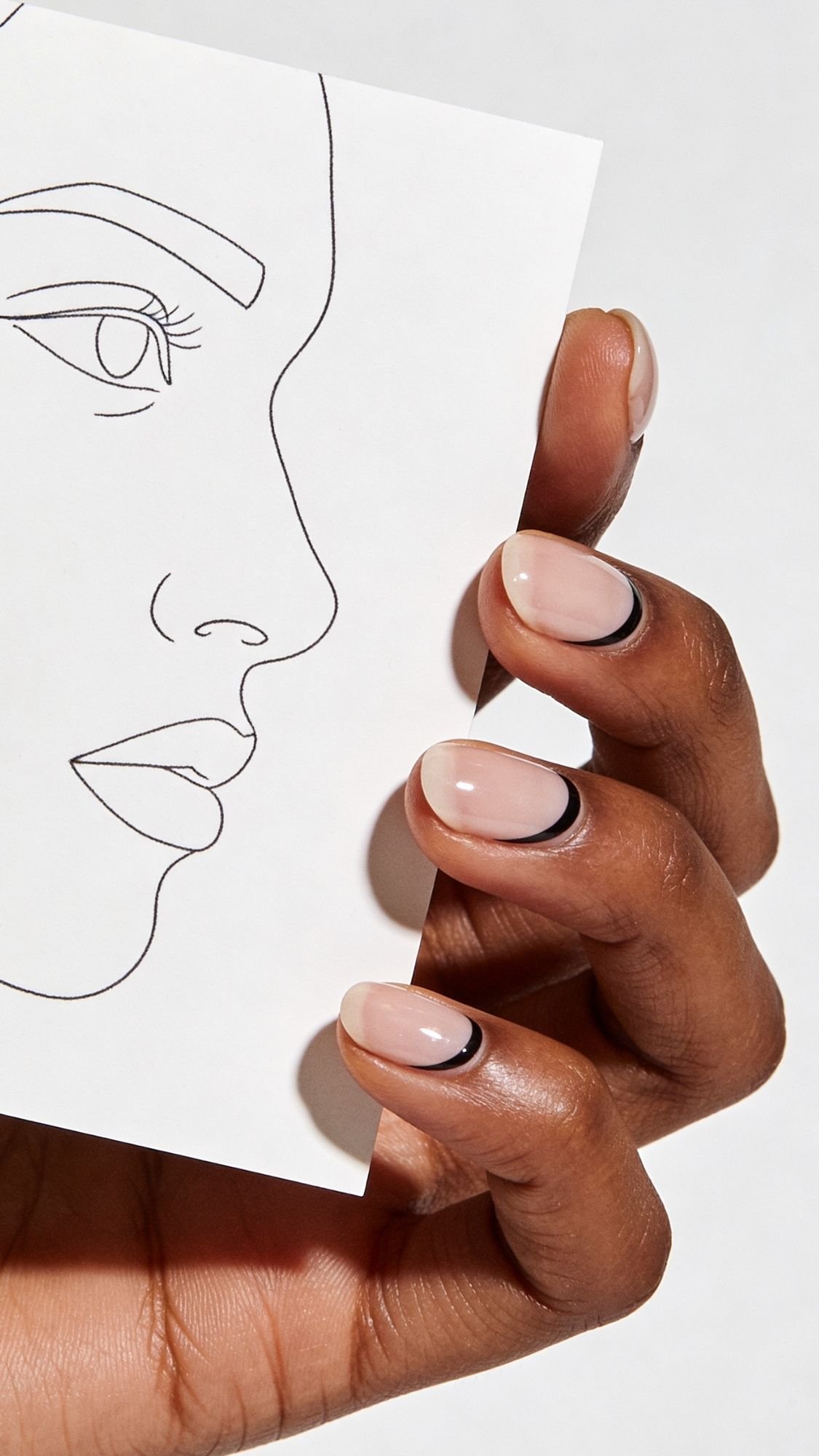 A hand with elegant, nude-colored nails and black outlines holds a white card featuring a minimalist line drawing of a woman's face in profile, offering inspiration for chic nail designs.