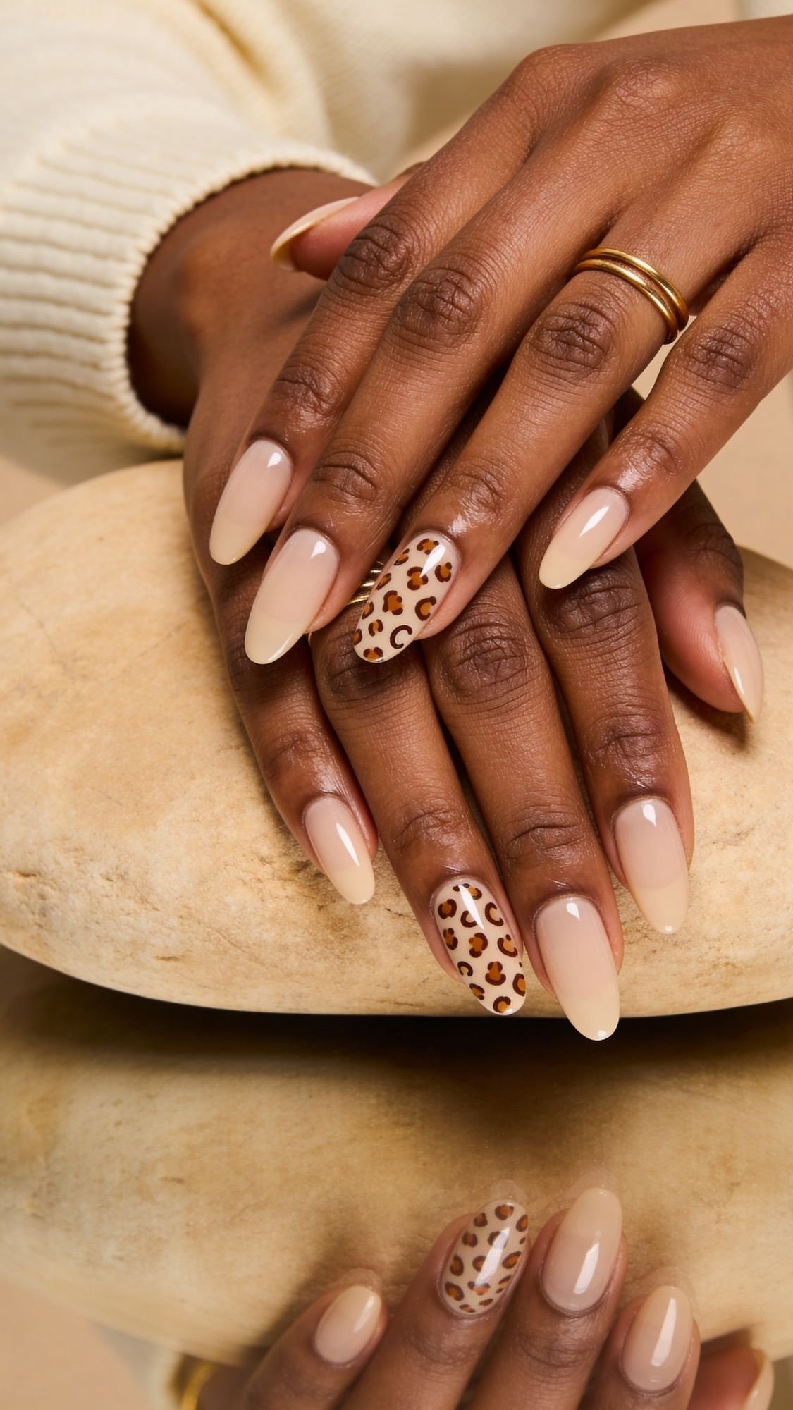 A close-up of hands with flattering almond nails in a nude shade; ring fingers feature a chic leopard print nail design. The hands rest on a smooth beige stone, with a gold ring and a cream sweater sleeve visible.