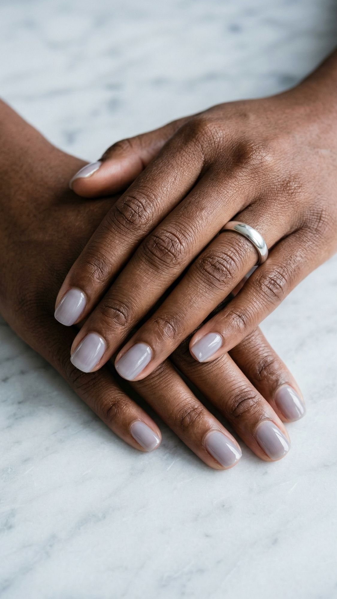 A close-up of two hands with dark skin, polished nails painted a pale lavender shade, and a simple silver band ring on the ring finger, resting on a light marble surface.