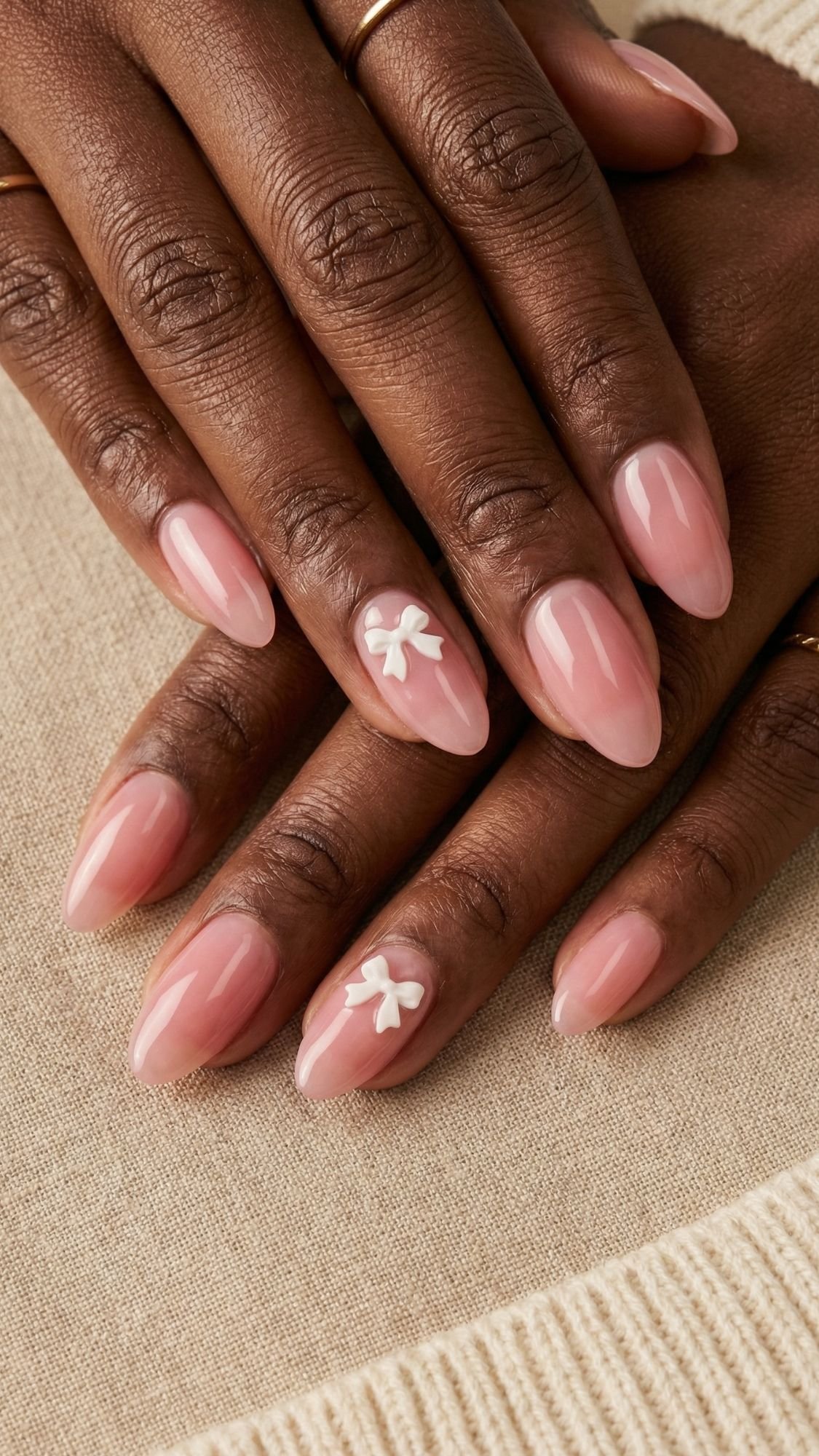 A close-up of hands with almond-shaped, glossy light pink nails. Two nails showcase charming 3D white bows, a trendy nail design. The person wears simple gold rings and a beige knit garment—perfect for those seeking flattering nails.