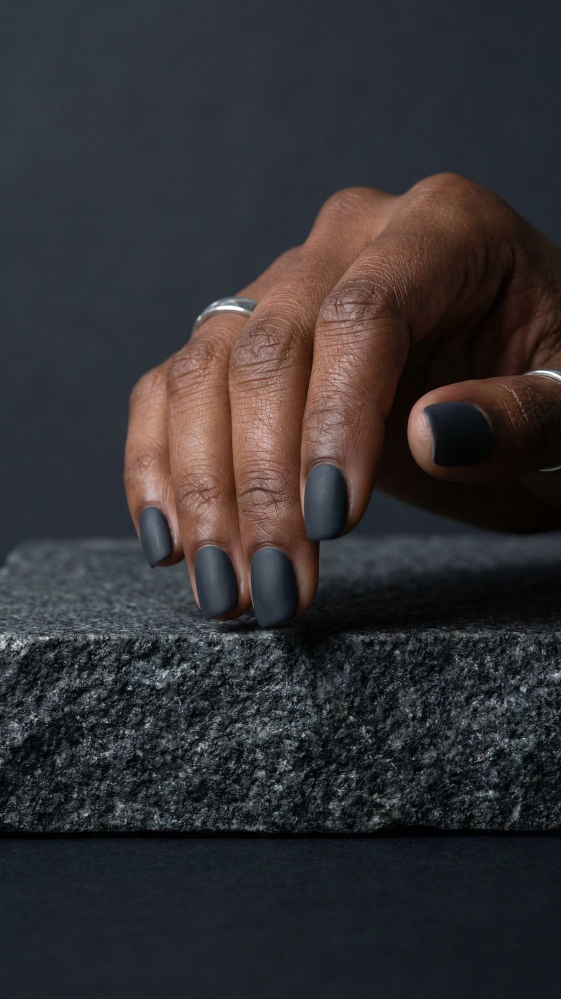 A hand with dark skin and matte dark gray polish shows off elegant nails, resting on a textured gray stone slab against a dark background. The short nails are adorned with simple silver rings.