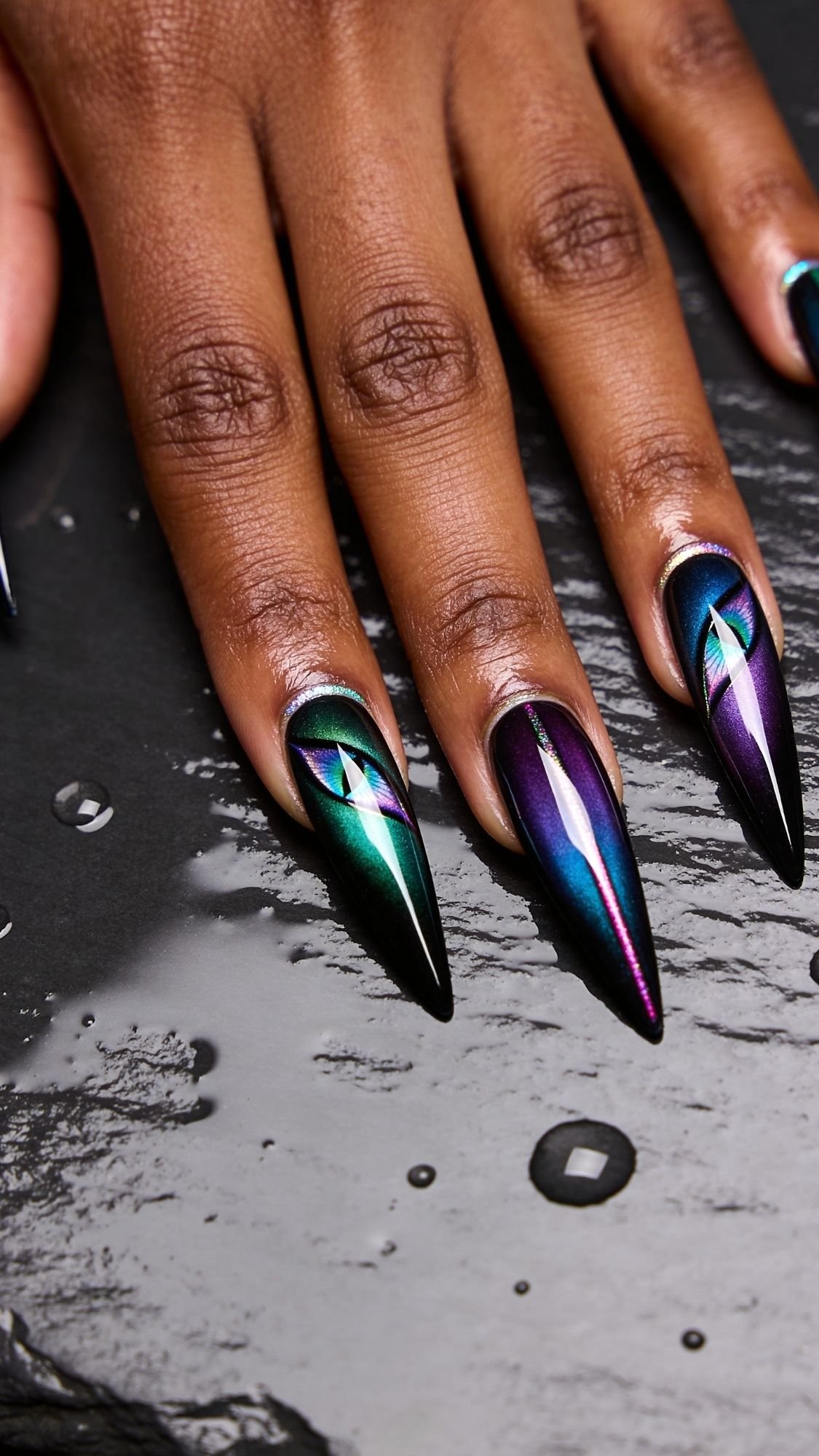 A hand with long, pointed nails featuring a glossy, geometric design in metallic green, blue, and purple shades rests on a black surface with scattered water droplets—perfect summer nails for any stylish brunch.