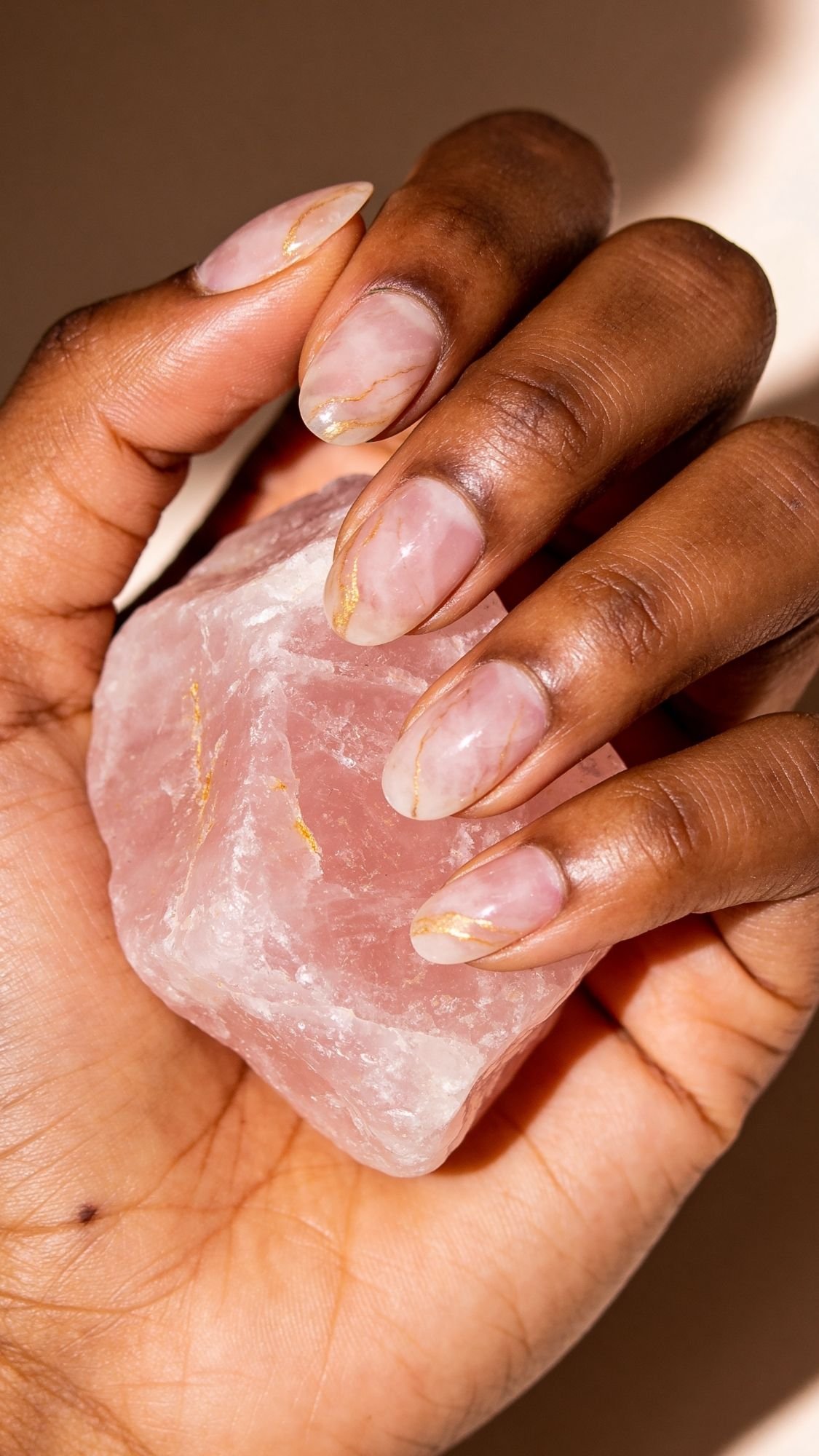 A hand with elegant, marble-patterned nude and gold manicured short nails holds a large, rough rose quartz crystal against a neutral background.
