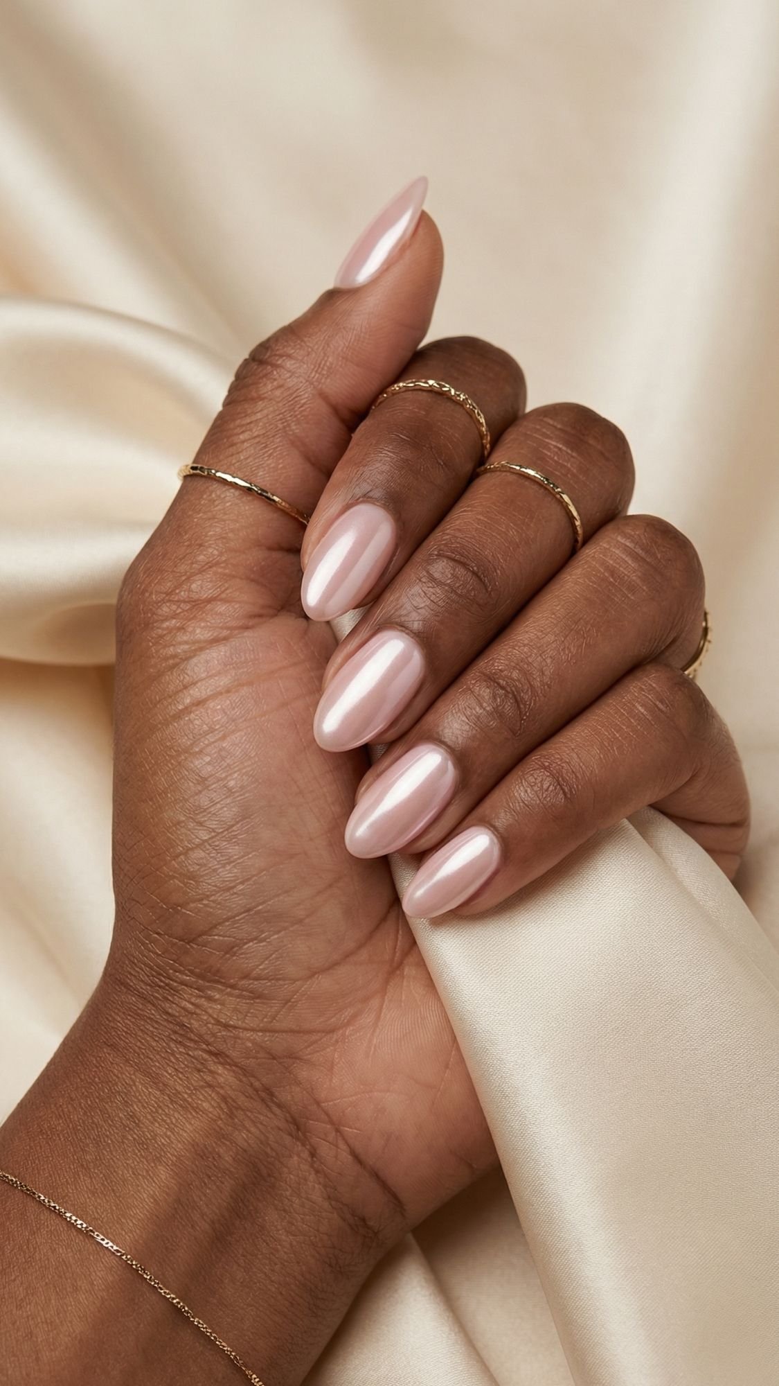 A hand with medium brown skin and almond-shaped, glossy pale pink nails holds cream-colored satin. Shiny nail designs catch the light, while delicate gold rings adorn several fingers for a chic selfie nails moment.