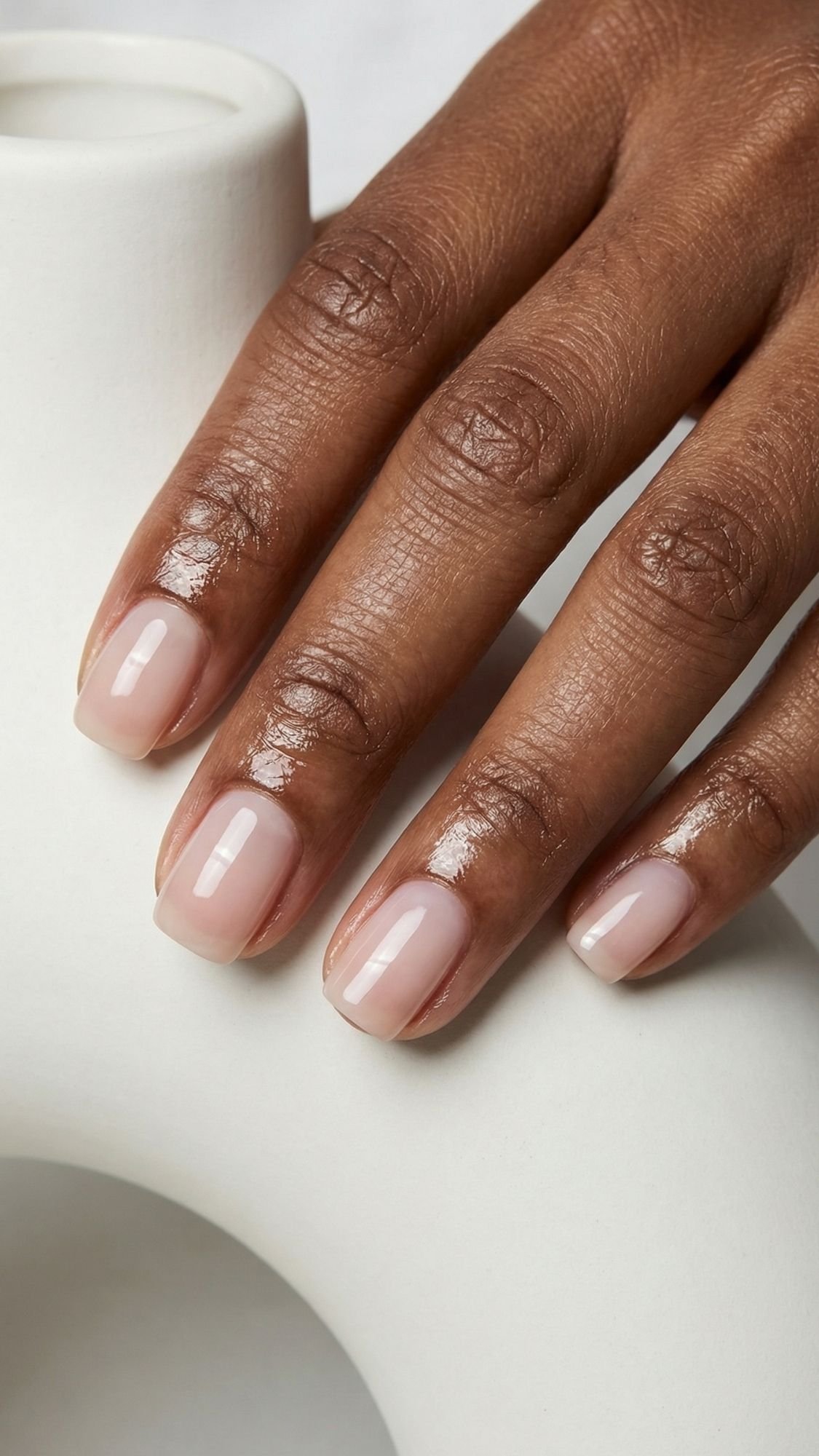 A close-up of a hand with dark skin showing neatly manicured, polished nails painted with a glossy, pale pink polish, resting on a smooth, white curved object—perfect for that clean girl nails look.