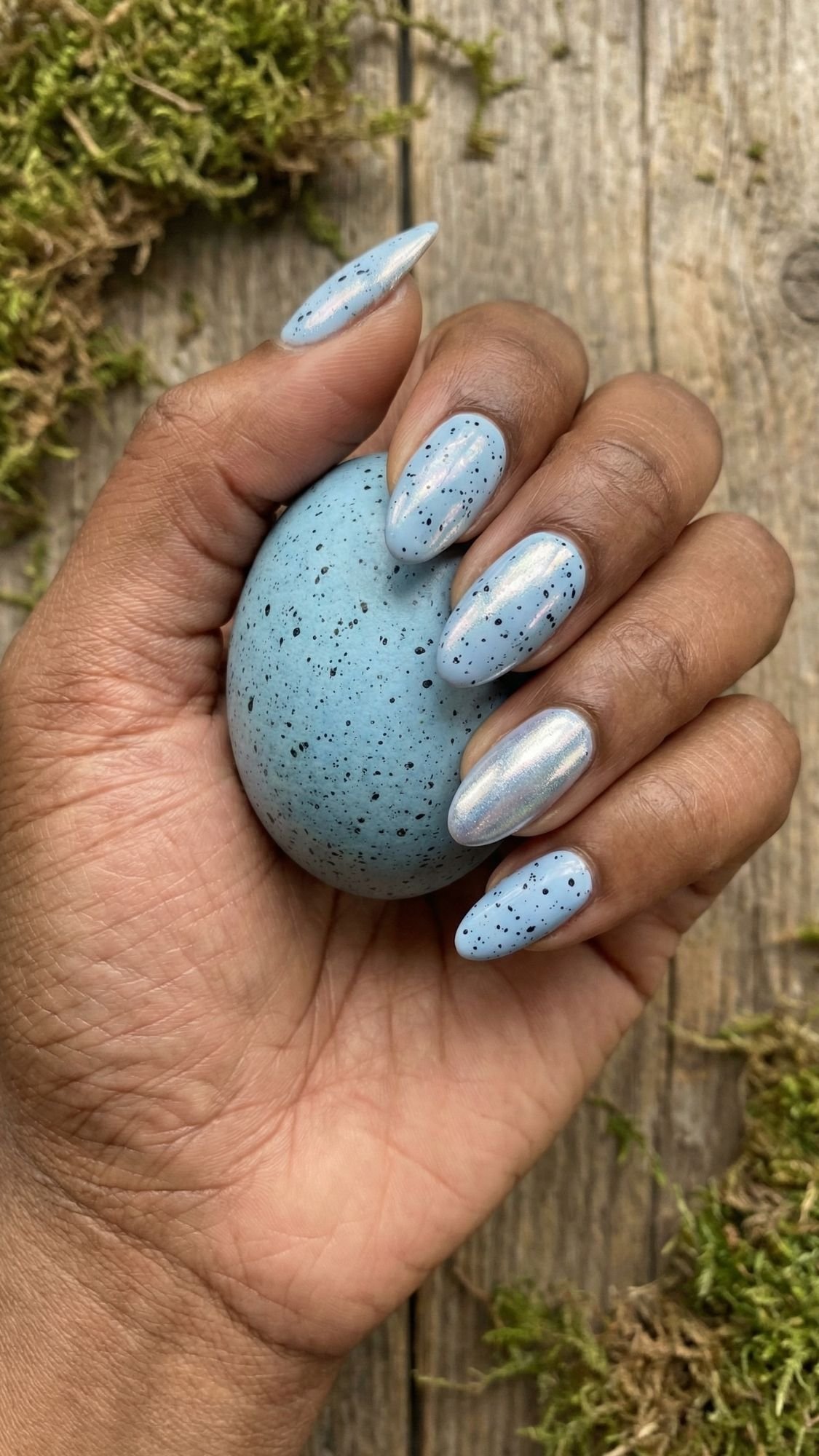 A hand with long, light blue and metallic silver holiday nails holds a blue speckled egg. The background is rustic wooden boards with patches of green moss.