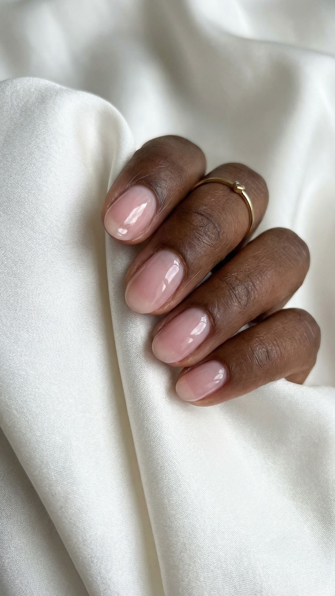A hand with neatly manicured short nails painted in a pale pink shade rests on white satin fabric. The person is wearing a thin gold ring on one finger, showcasing elegant nails and effortless style.
