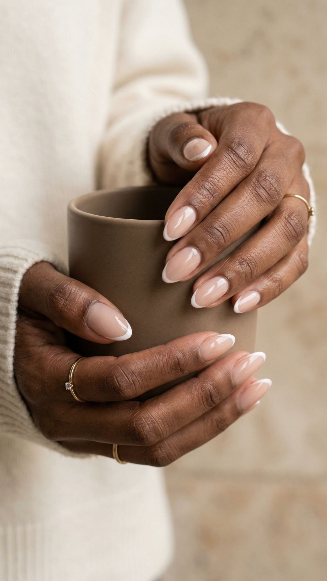 A person with neatly manicured, almond nails featuring nude and white French tip nail designs holds a matte brown mug. They wear a cream-colored sweater and thin gold rings on their fingers.