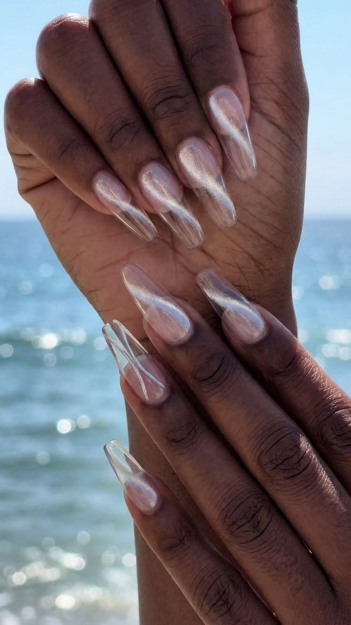 Two hands with long, almond-shaped nails painted with a shiny, iridescent nude polish are held in front of a blurred ocean background under daylight—perfect beach nails for your next summer outing.