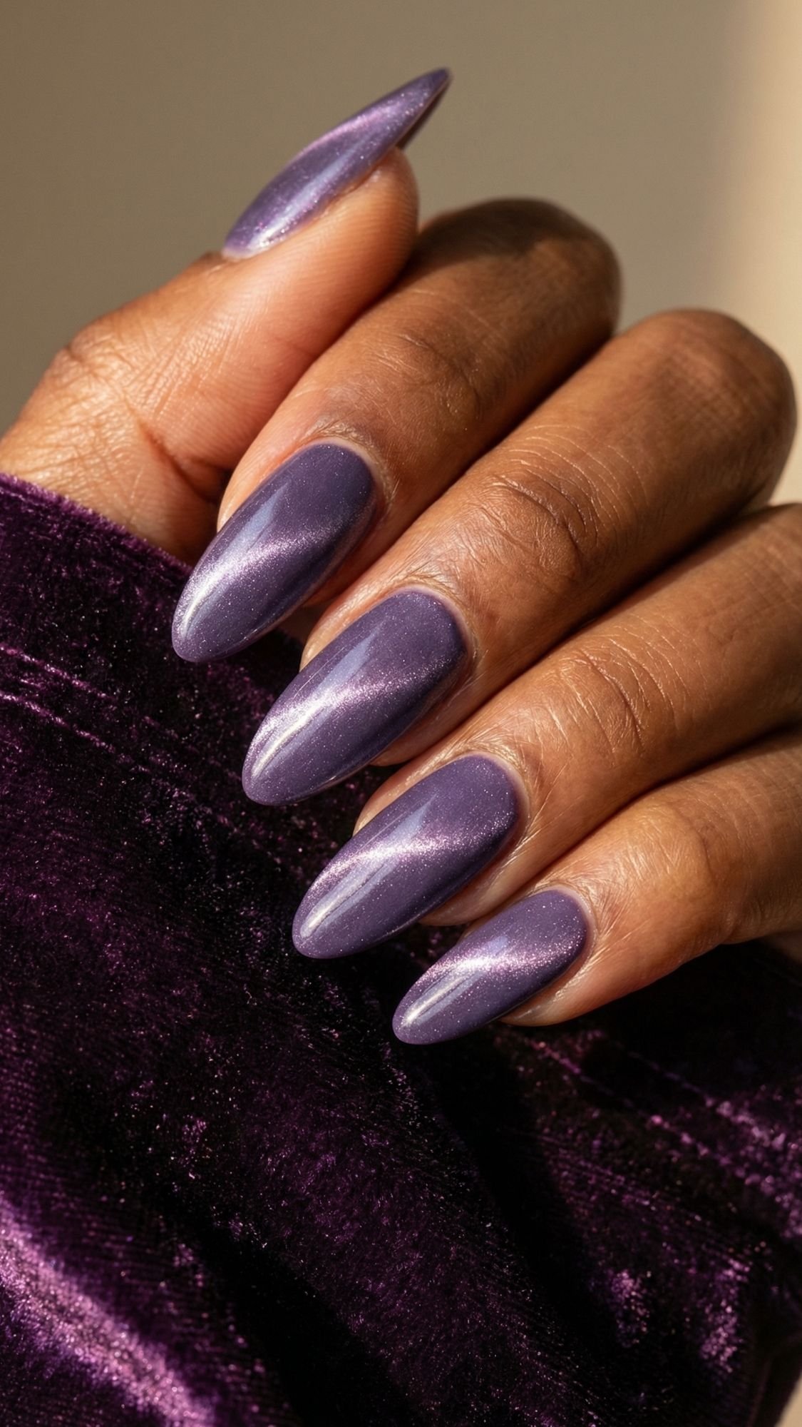 A close-up of a hand with long, almond-shaped nails painted in metallic purple polish with a hint of sparkle. The person is wearing a deep purple velvet sleeve, perfect for adding glamour to any special occasion.