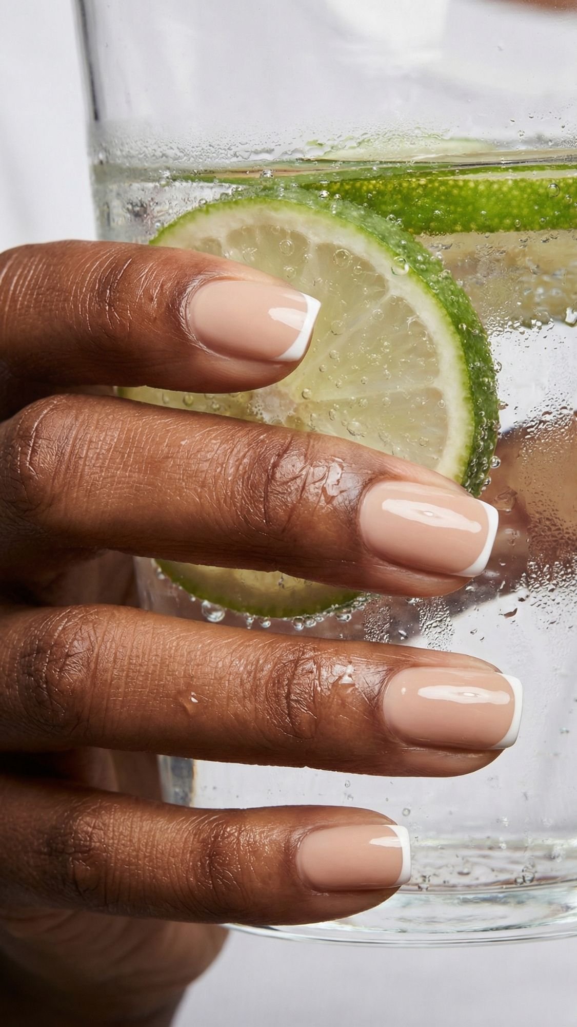 A hand with neatly manicured, elegant nails featuring a nude base and white tips holds a glass of sparkling water with lime slices inside.