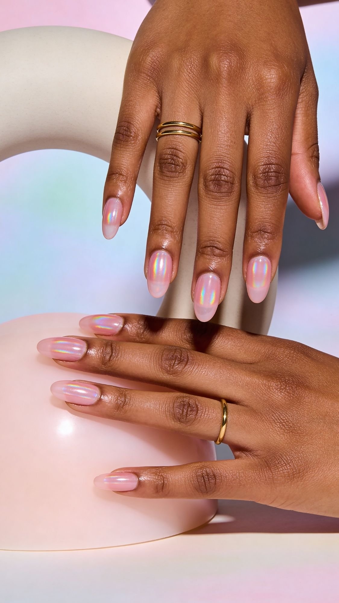 Two hands with gold rings display manicured nails painted with shiny, iridescent chrome nails. The selfie showcases the hands against a soft, pastel-colored background with smooth, curved props.