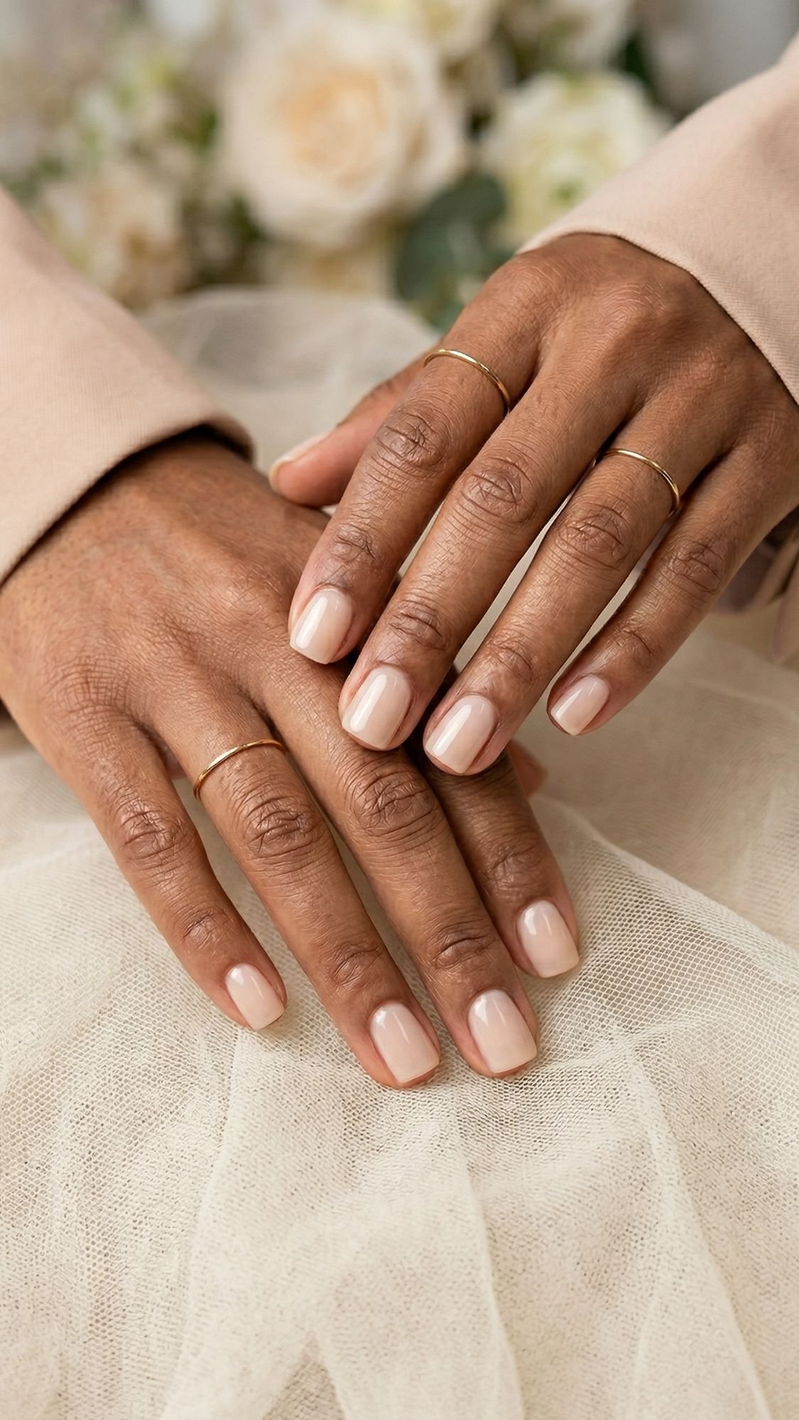 Two hands with neatly manicured nails painted in a light nude color rest gently atop each other—perfect wedding guest nails. The person wears thin gold rings on several fingers, and a soft, neutral fabric is in the background.