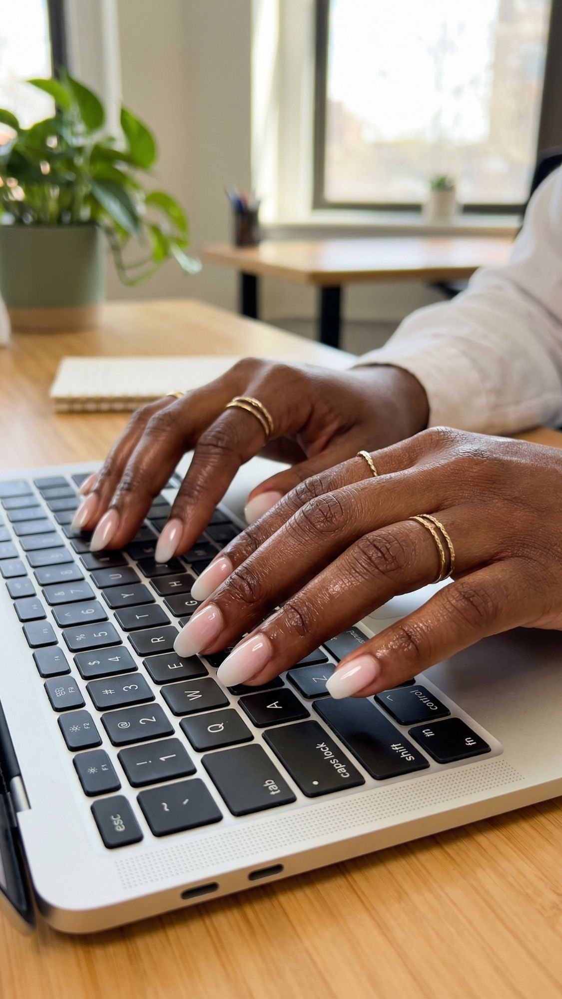 A person with chic, work friendly nails and gold rings types on a laptop keyboard at a wooden desk, with a notepad, pen, and potted plant visible in the background.