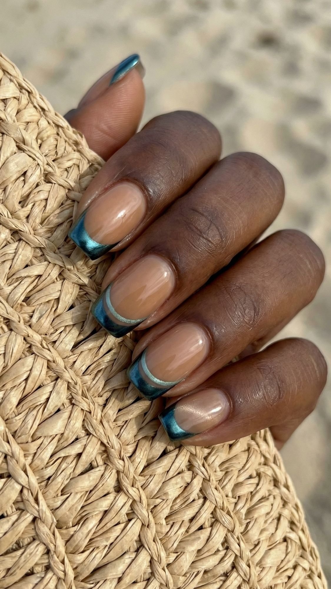 A hand with short, natural-shaped nails features a glossy nude base and teal and silver double French tips—perfect summer nails. The hand is holding a woven straw textured item, with a sandy background visible.