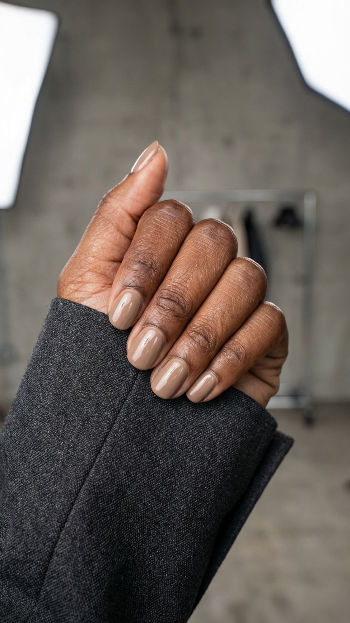 A close-up of a hand with medium brown skin, showing neatly manicured chic nails painted in a glossy beige color. The person is wearing a dark gray sleeve. The background is blurred and industrial-looking—perfect for work friendly nails.