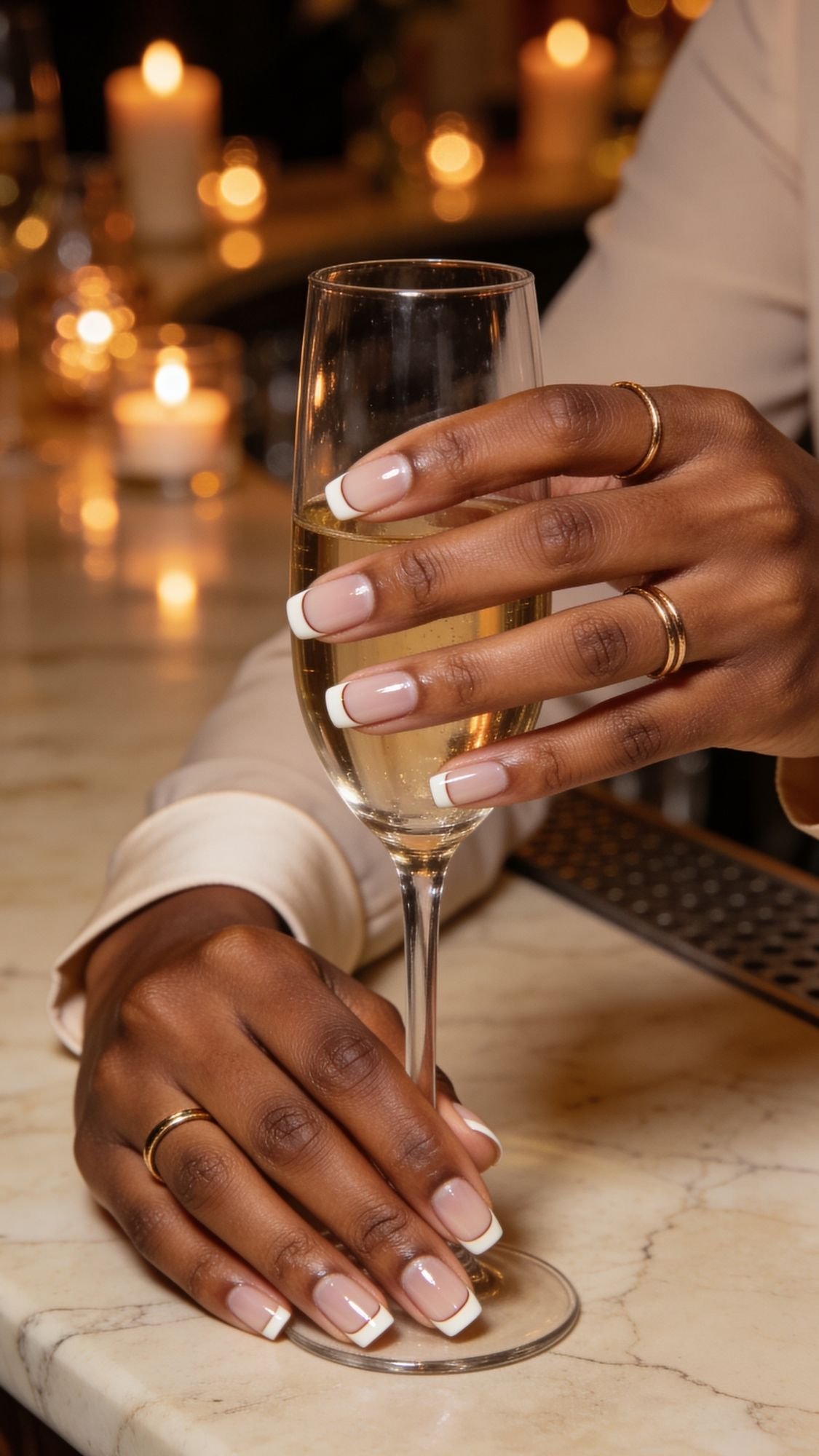 A person with neatly manicured French-tip nails for wedding and gold rings holds a champagne glass on a marble countertop, with softly lit candles in the background.