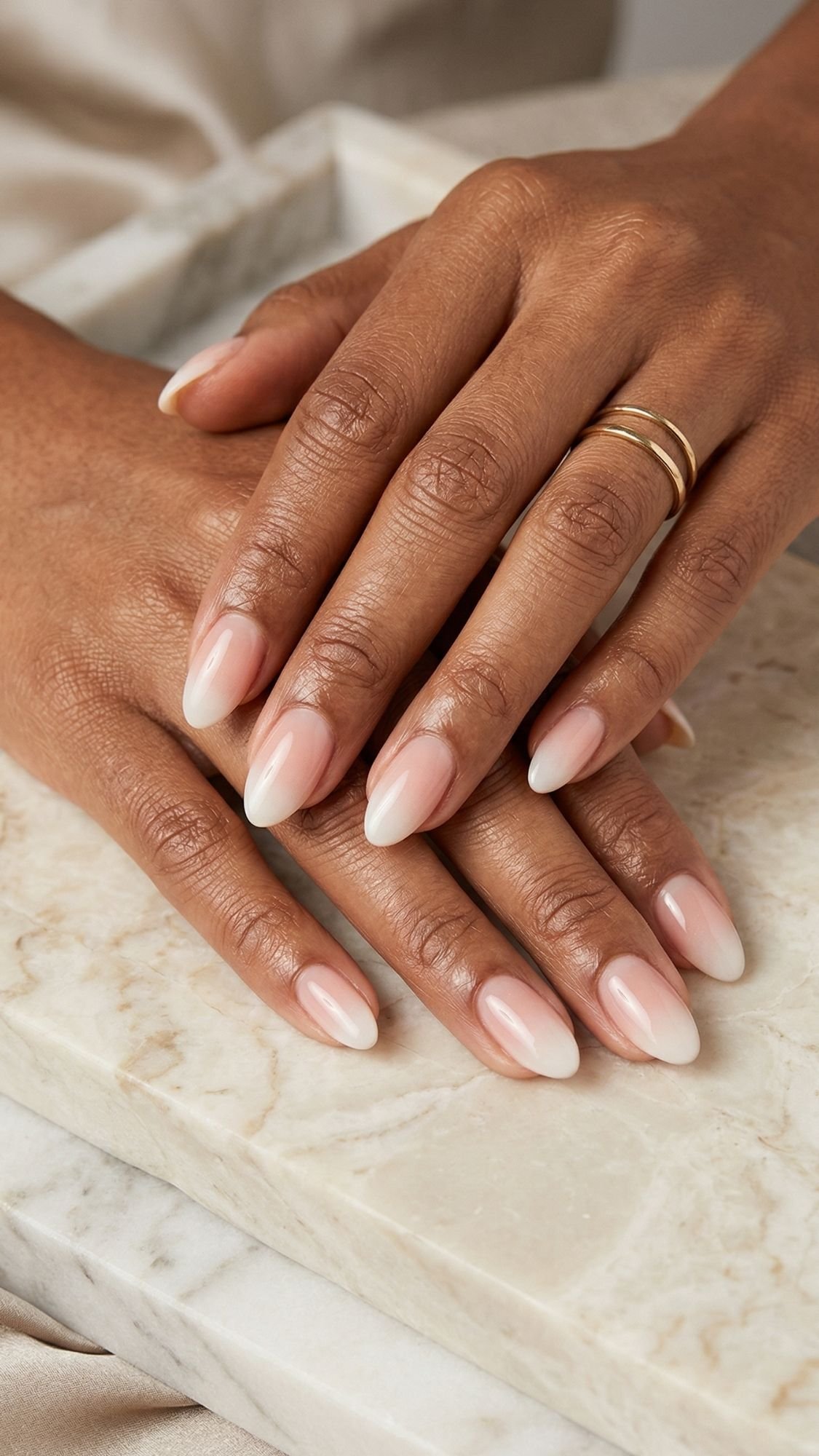 Close-up of two hands with flattering almond nails, neatly manicured and painted in a glossy, natural pink shade, resting on a light marble surface; one hand wears a simple gold ring, showcasing elegant nail designs.