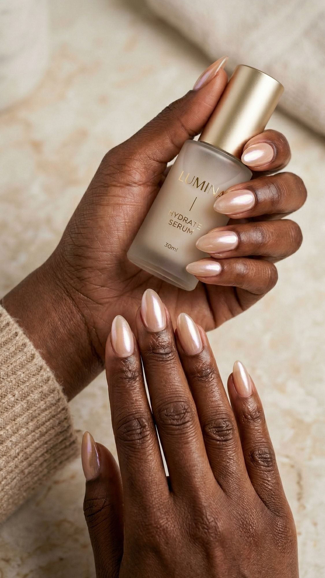 A person with neatly manicured, almond nails in a pearly nude shade holds a bottle of facial serum labeled Hydra Serum in one hand, while the other hand rests flat on a marble surface, showcasing one of the most flattering nail shapes.