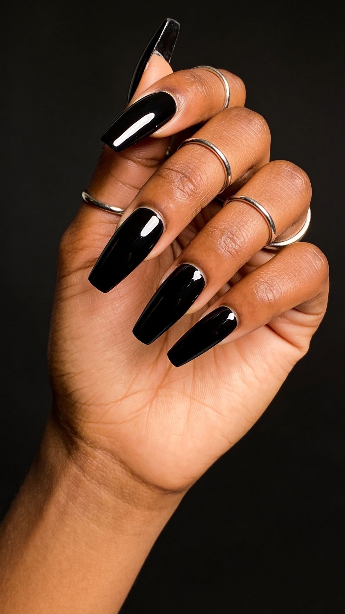 A hand with long, glossy black coffin-shaped nails and multiple silver rings on each finger, posed against a black background, showcases striking light catching nails.