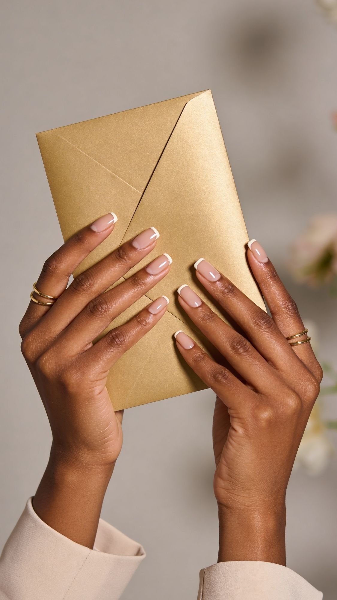 A person with neatly manicured French-tip nails—perfect wedding guest nails—and gold rings on their fingers holds a golden envelope against a blurred neutral background.