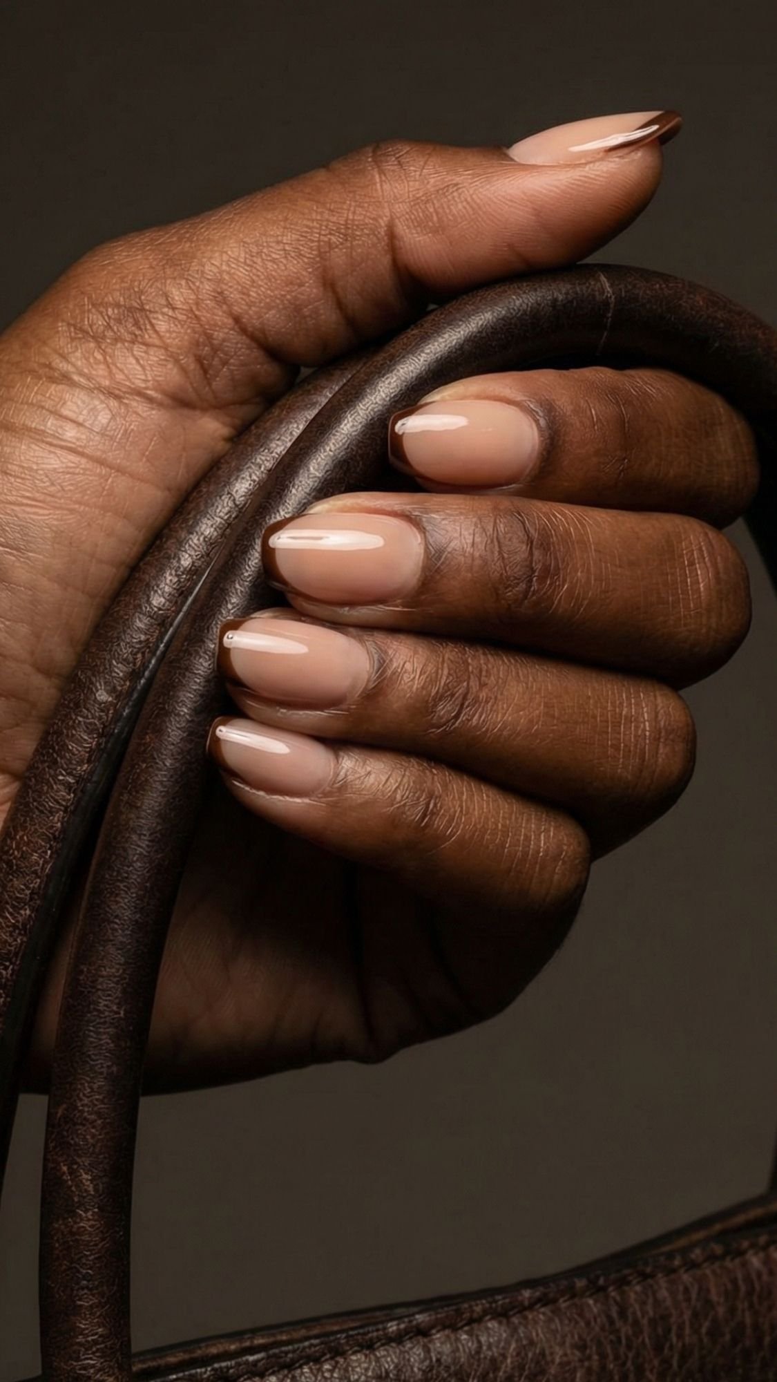 A hand with medium brown skin holds a dark brown leather strap. The short nails are manicured with glossy nude polish for an elegant nails look. The background is dark and neutral.